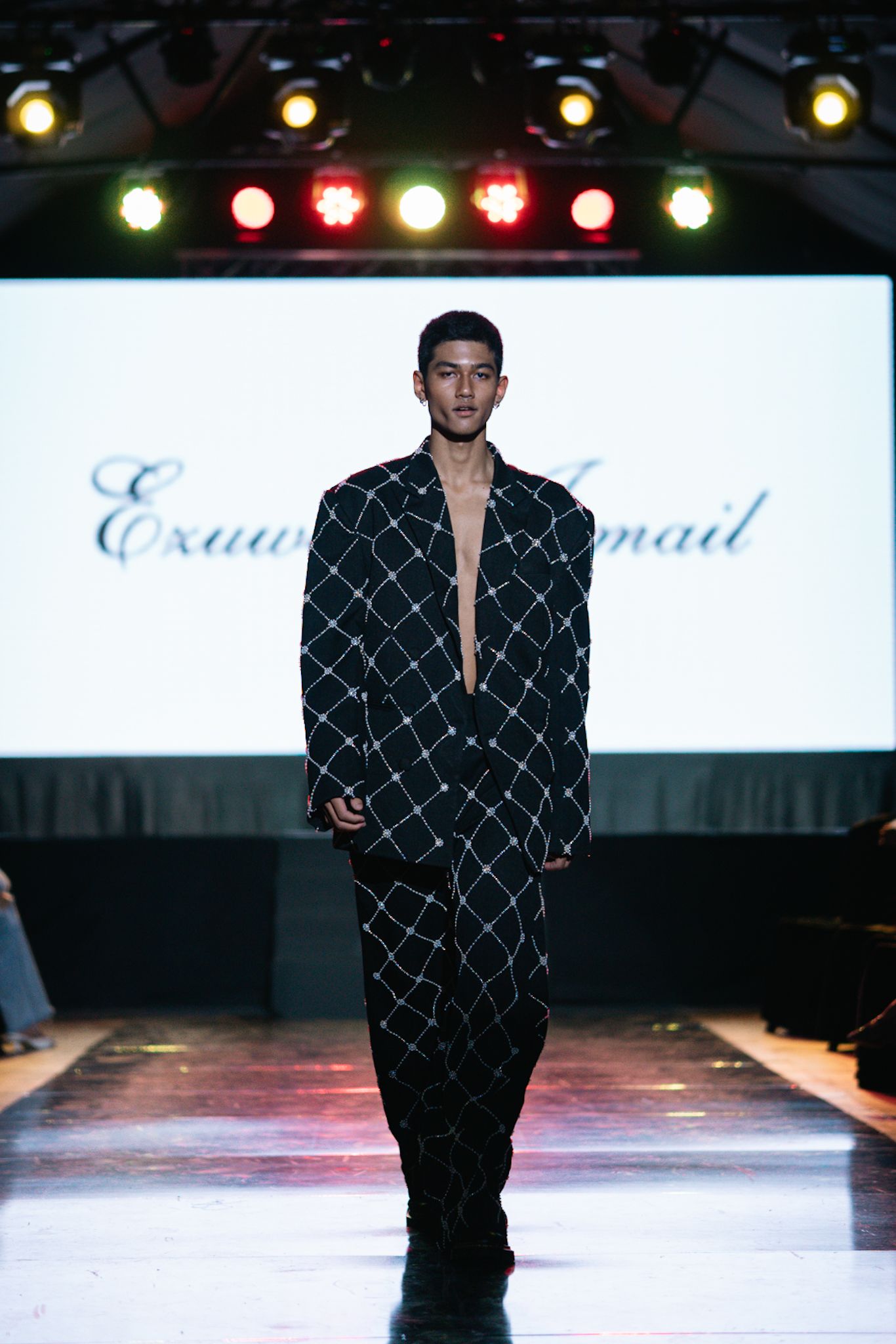 Model showcases black geometric patterned suit on illuminated fashion runway with stage lighting.