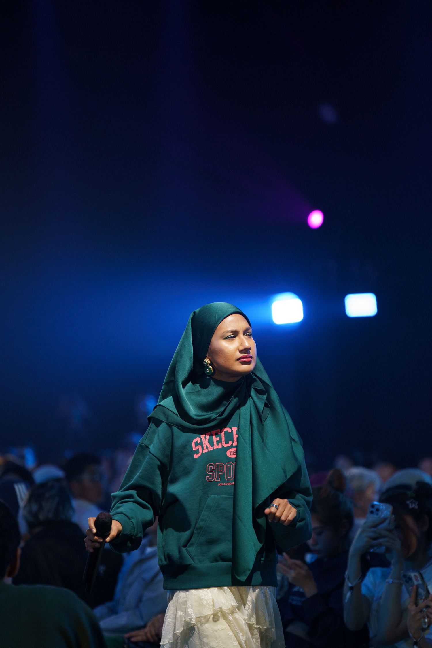 A person in a green hoodie and hijab stands against a dark blue backdrop with stage lighting.