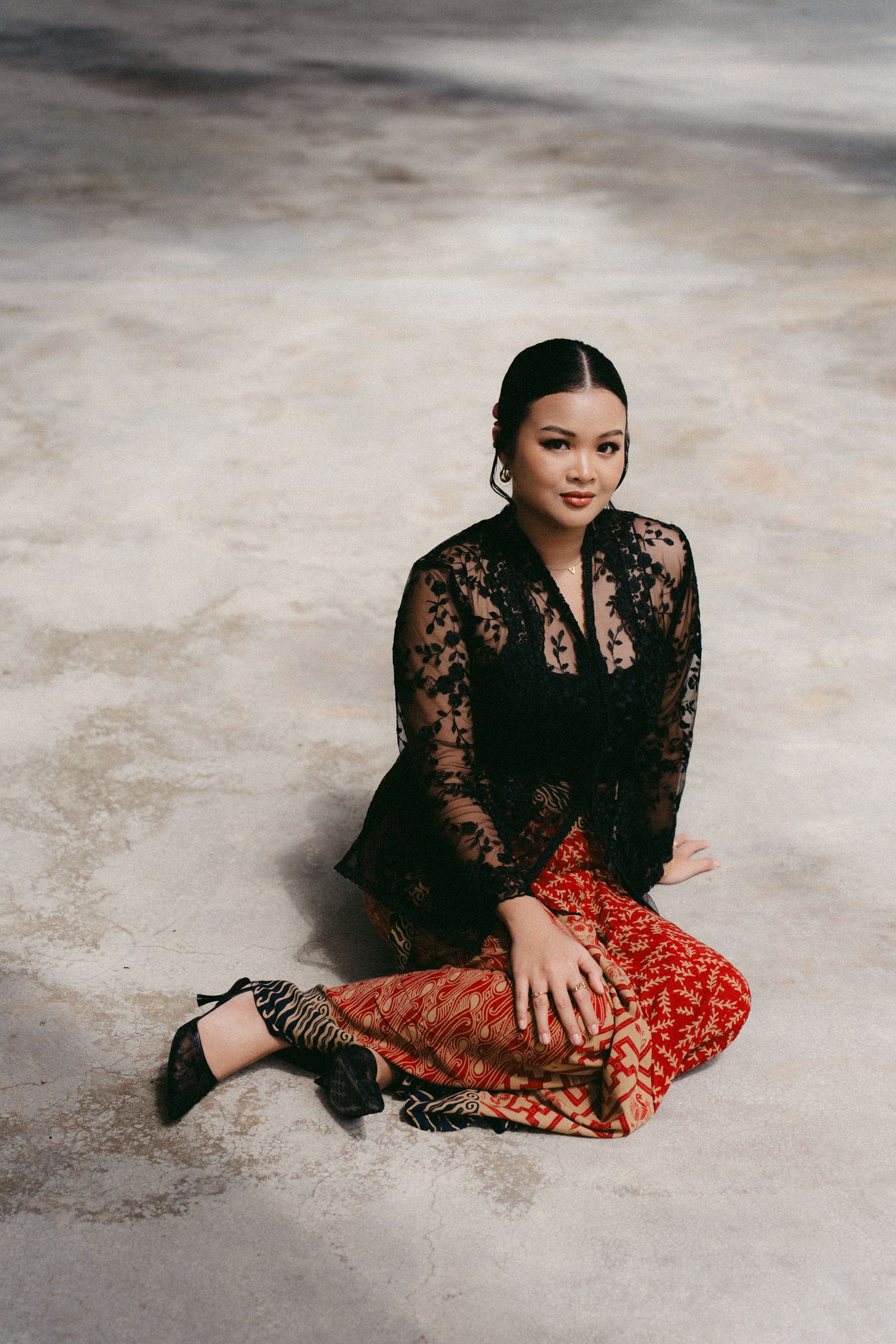 Person in black lace top and red patterned skirt sits elegantly on concrete floor during fashion photoshoot.