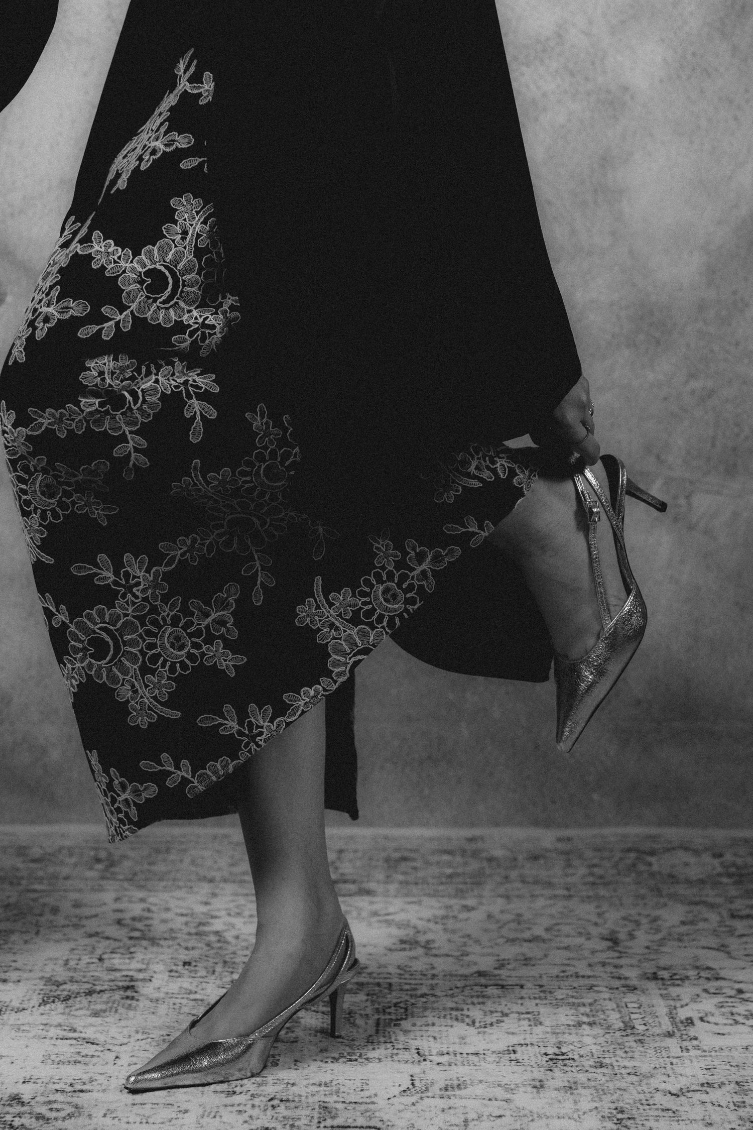 Black dress with intricate floral embroidery and elegant kitten heel shoes shown in dramatic black and white photography.
