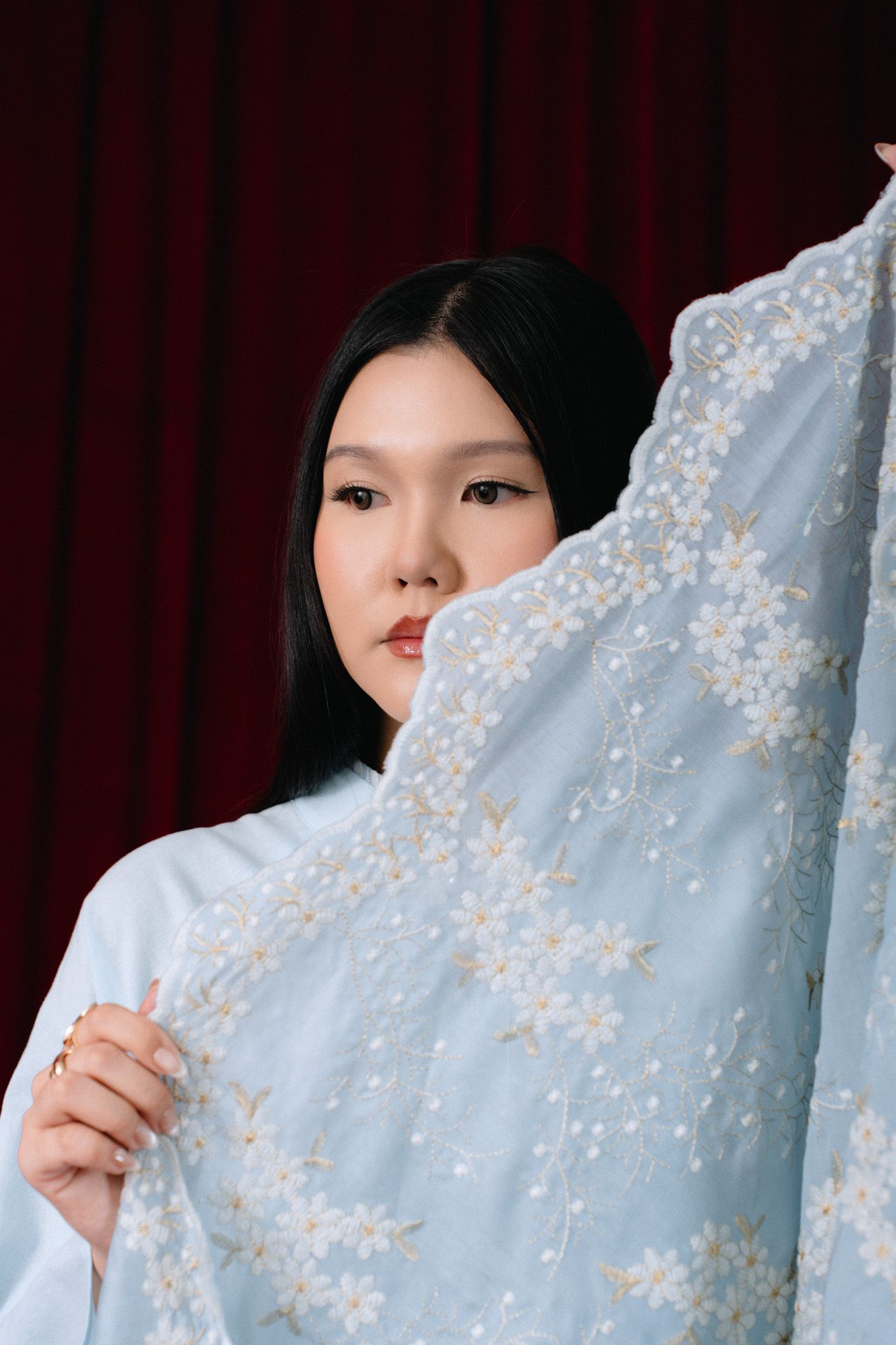 Asian woman in light blue floral embroidered dress holding up delicate fabric against red curtain backdrop.