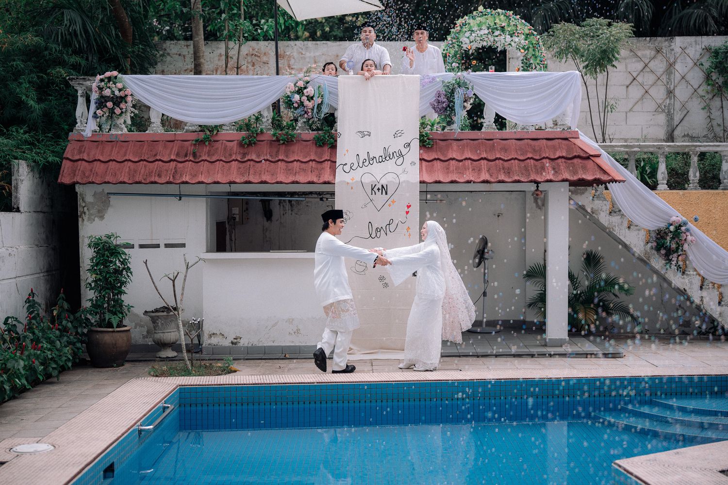 Couple in white attire dancing by a pool at an outdoor wedding celebration with tropical backdrop.