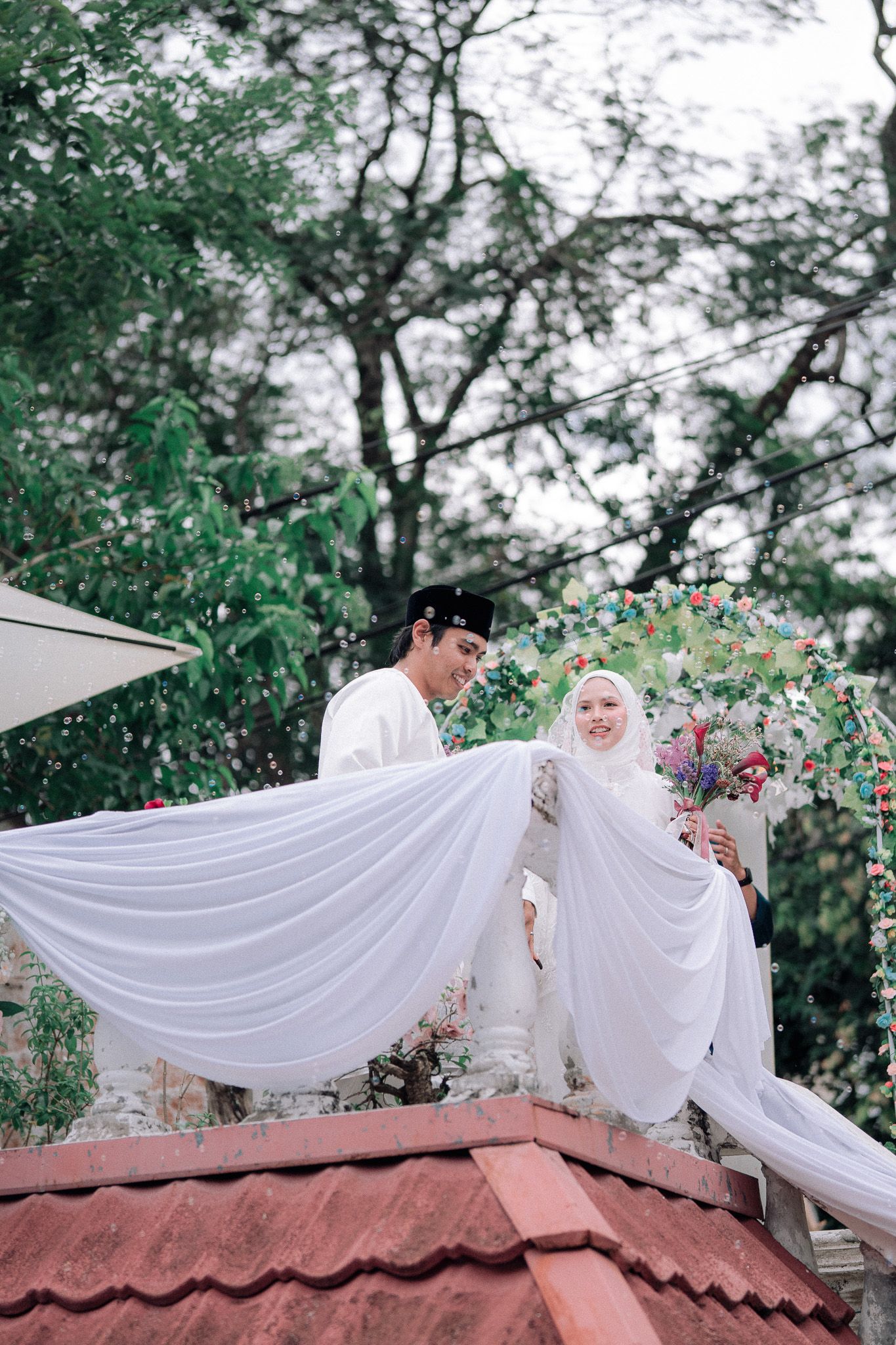 Muslim couple in white wedding attire pose outdoors, bride in hijab holds bouquet beside groom in traditional songkok cap.