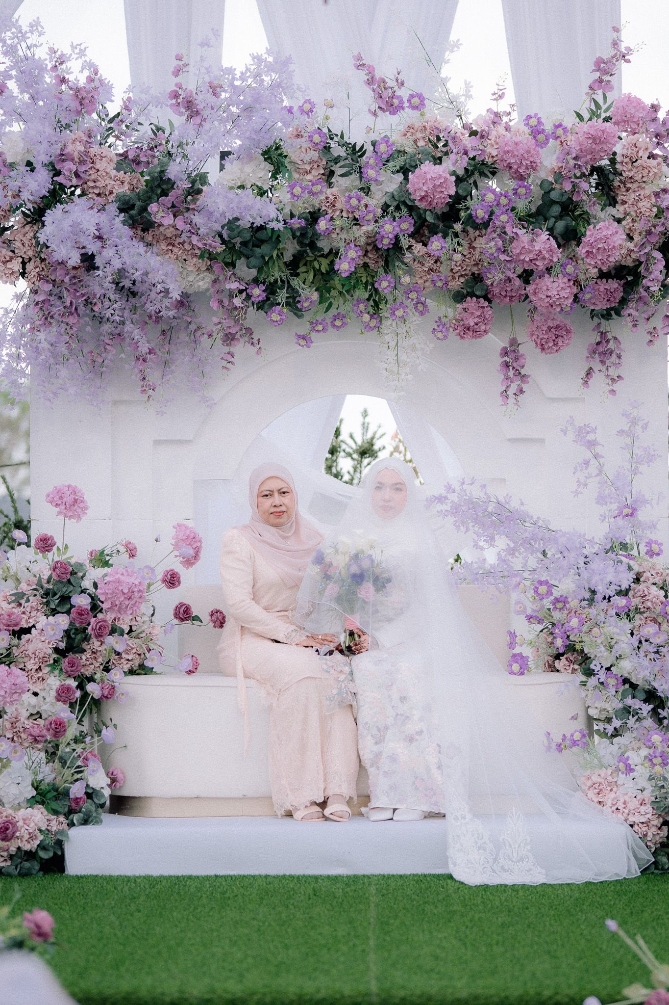 Two women in hijabs sit under a floral arch with purple and pink blooms, one in a white bridal gown.