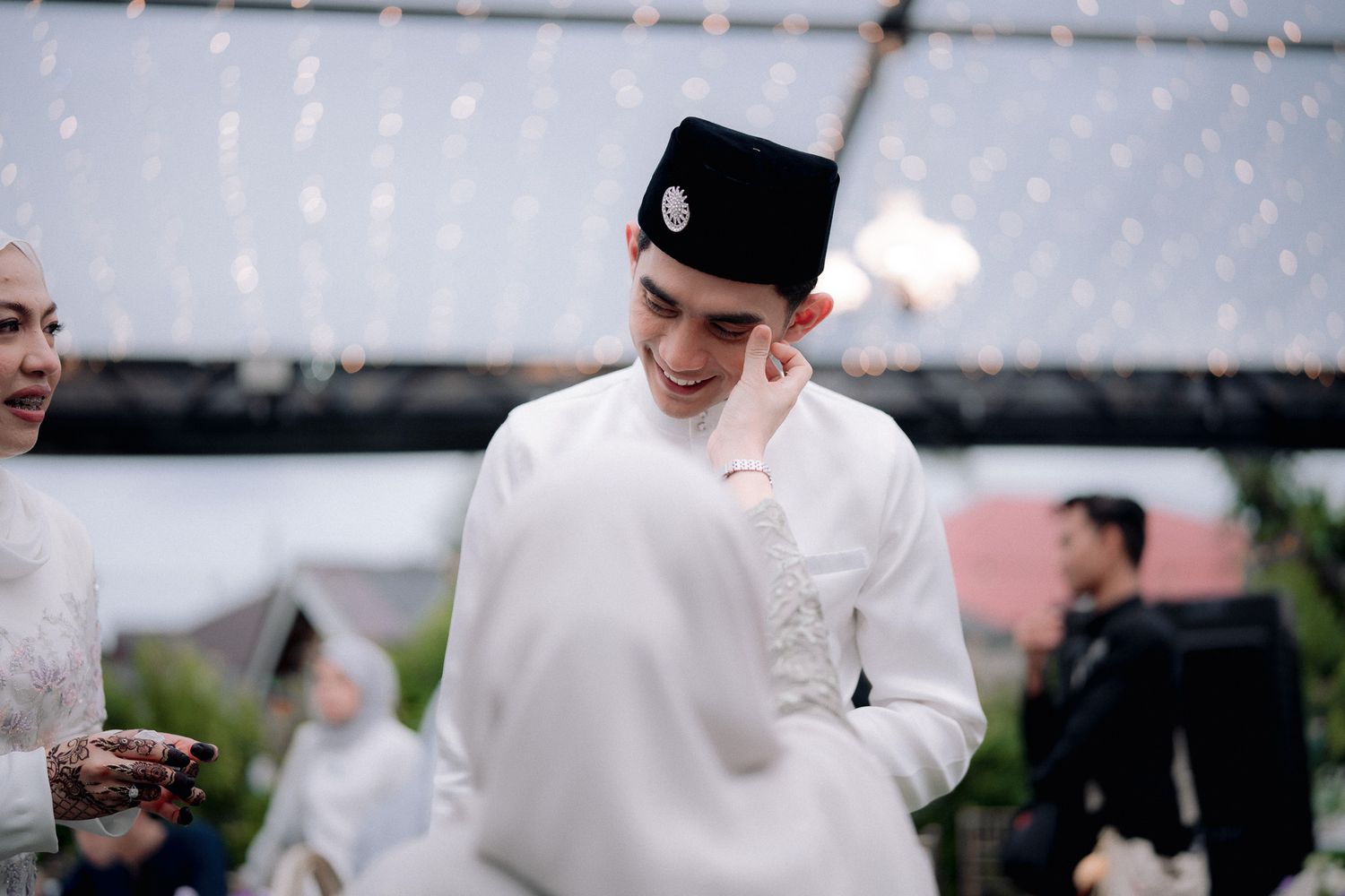 Young man in white baju melayu and songkok smiling shyly at a Malay wedding ceremony outdoors.