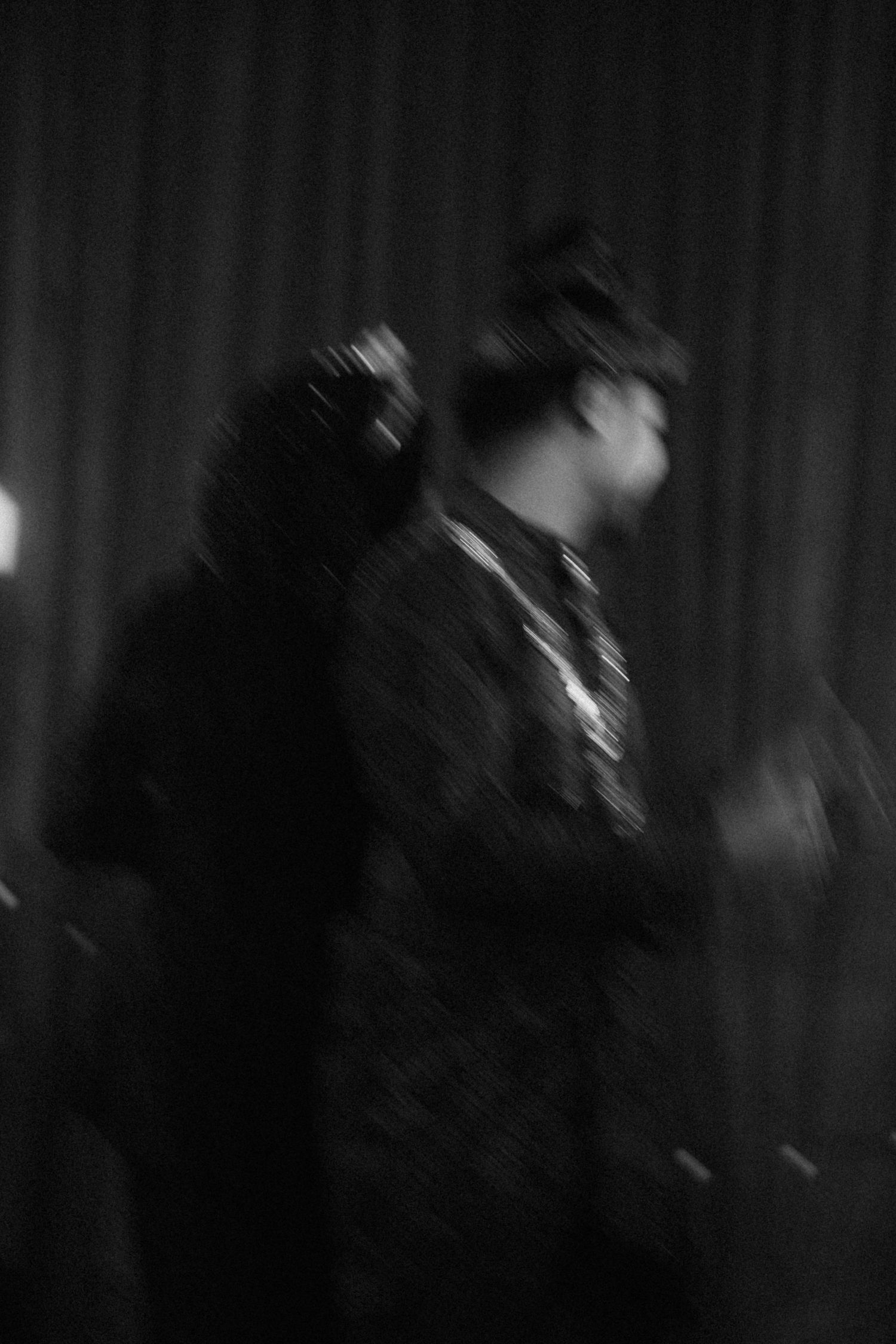 Blurry black and white artistic photograph showing a figure in motion wearing a hat and jacket against a dark background.