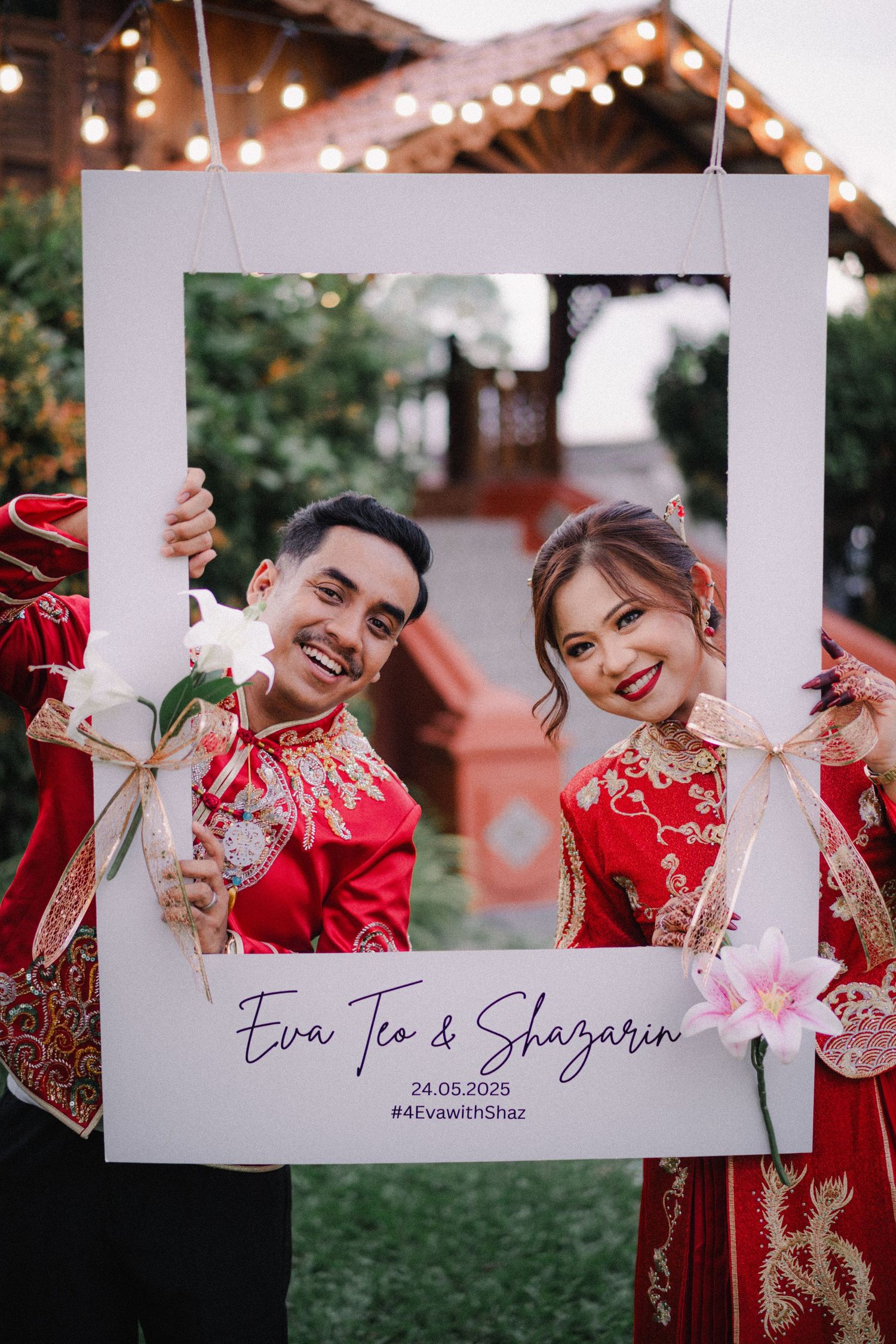Couple in traditional red Chinese wedding attire posing with a decorative white photo frame under string lights outdoors.