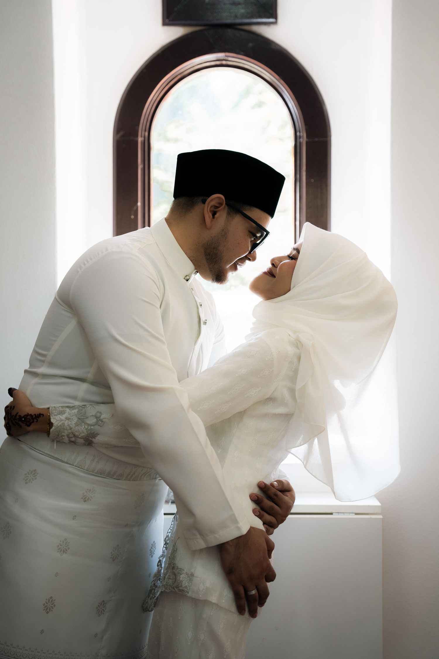 A tender moment between a couple in traditional Muslim wedding attire standing in front of an arched window.