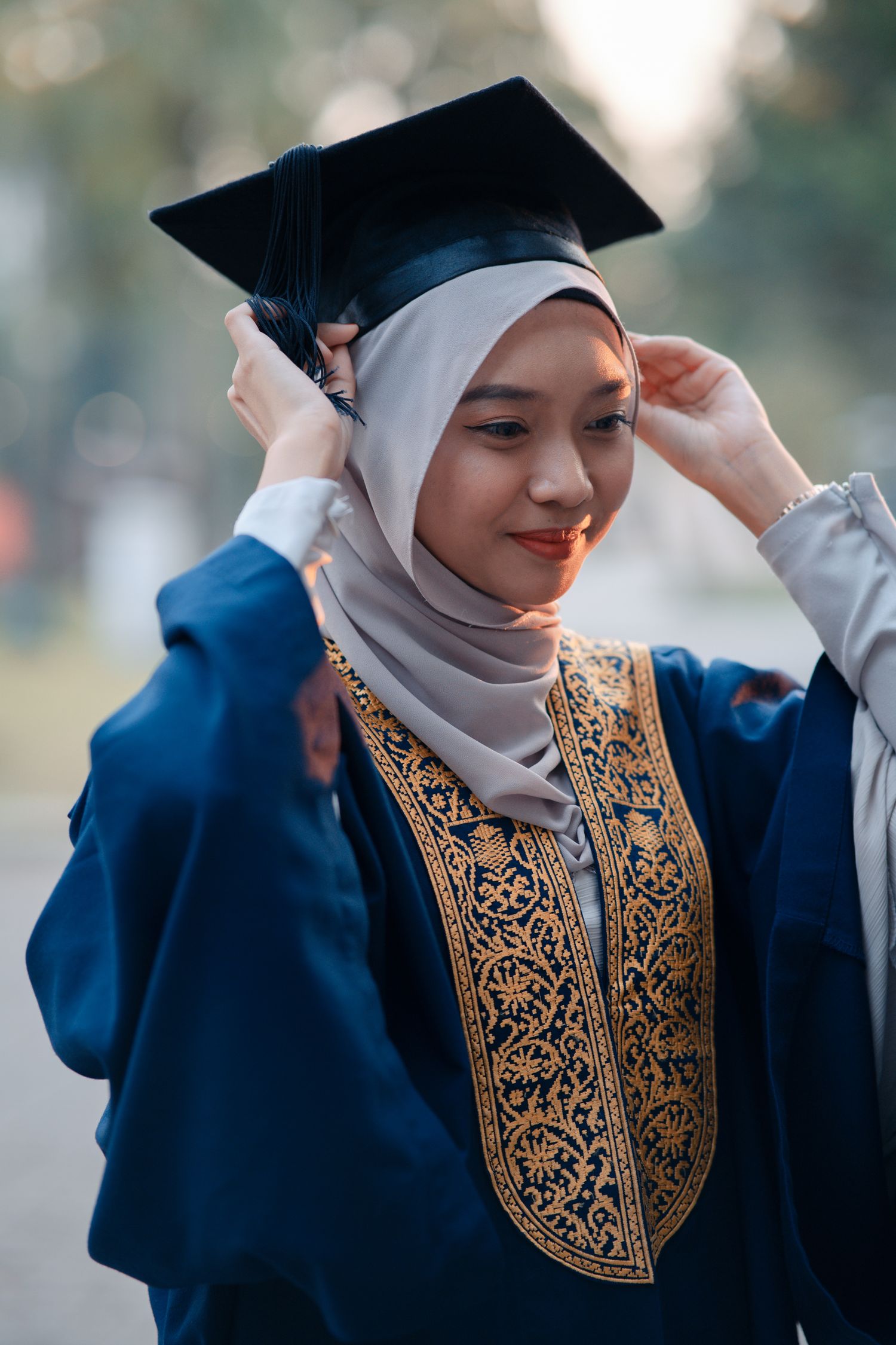 Graduate in blue gown wearing embroidered traditional dress adjusting cap.