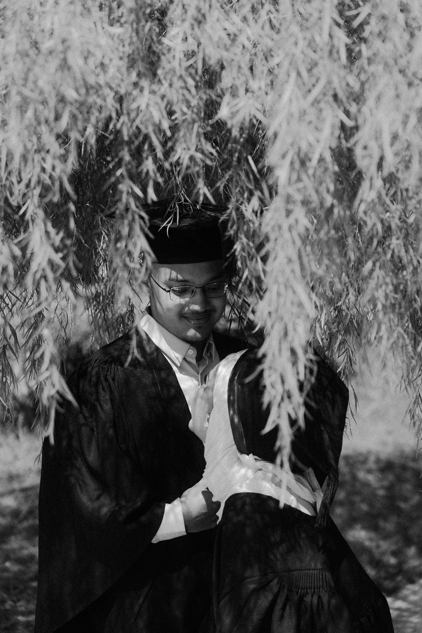 Black and white photo of a graduate in cap and gown holding diploma under weeping willow tree.