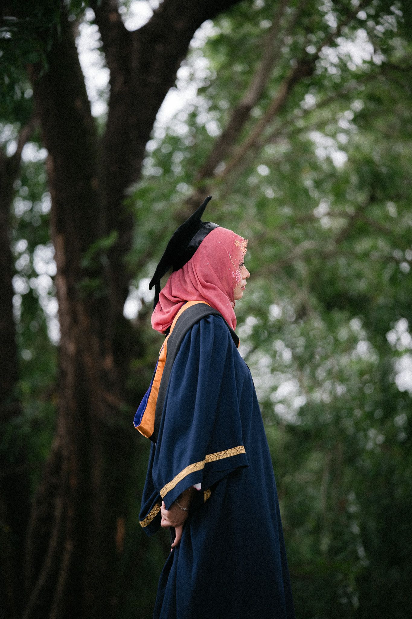 A Muslim woman in graduation gown, mortarboard cap, and pink hijab stands outdoors among green trees.