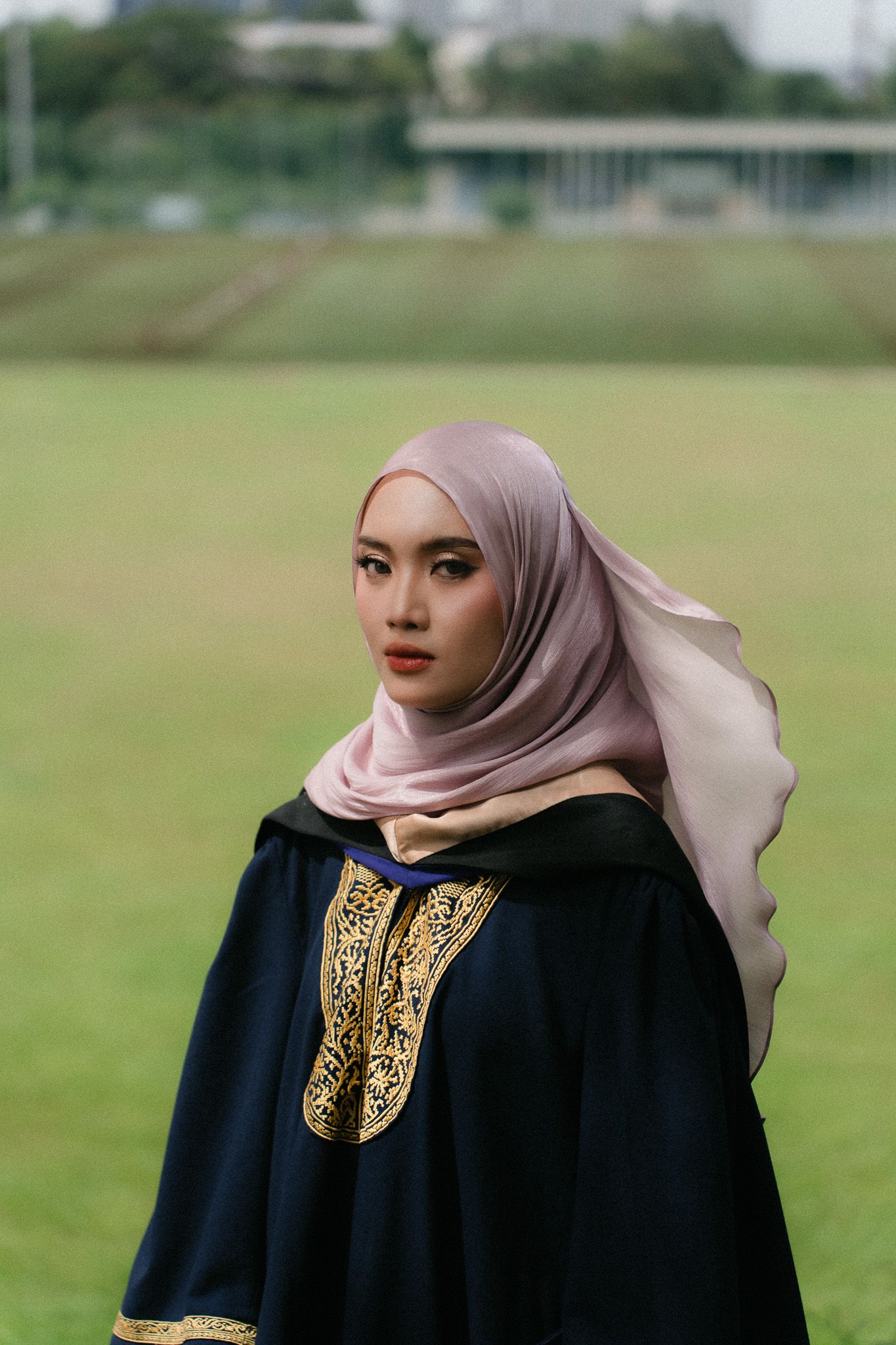 Young Muslim woman in black abaya with gold embroidery and pink hijab standing outdoors on green field.