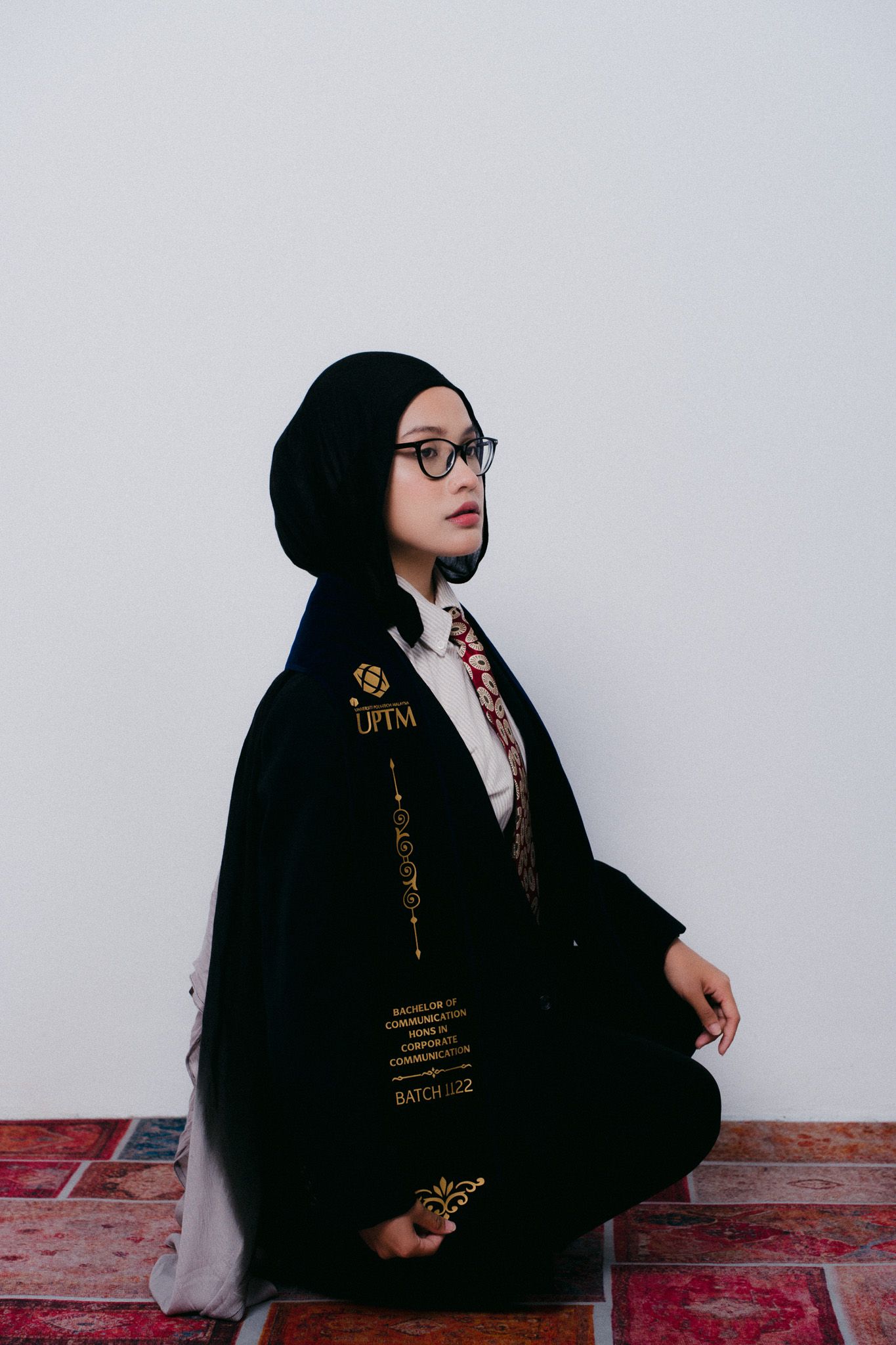 Young woman in black graduation gown and hijab with glasses sits gracefully on patterned floor against white wall.