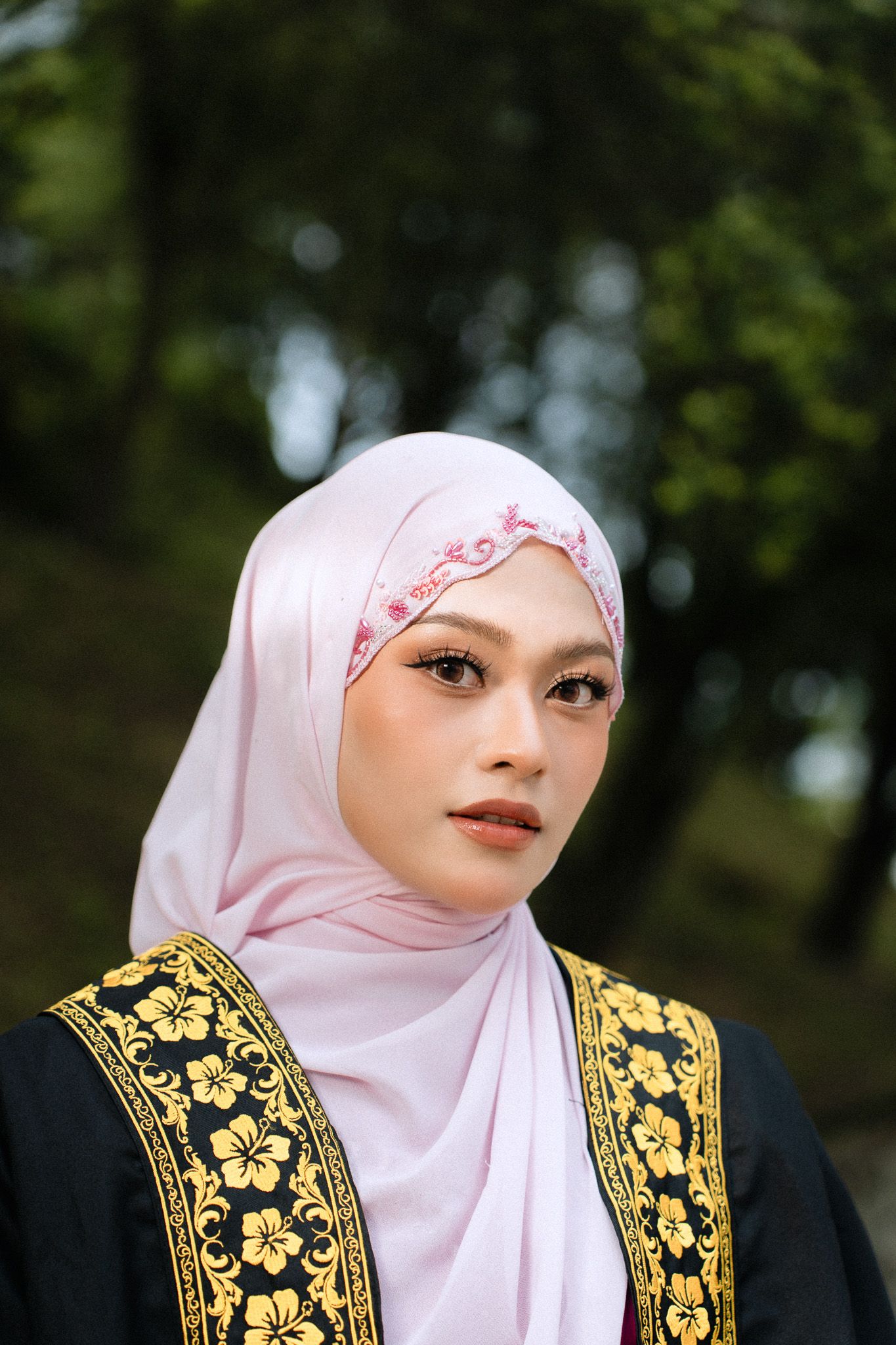 Young Muslim woman in pink hijab with floral embroidery and black gold traditional outfit posing outdoors.