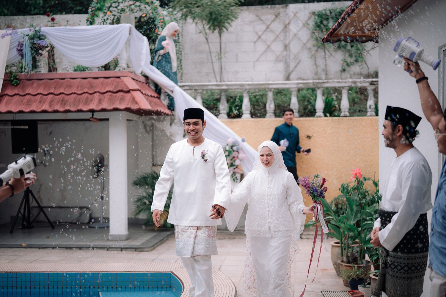 Muslim couple in white wedding attire holding hands walking past pool at outdoor garden ceremony.