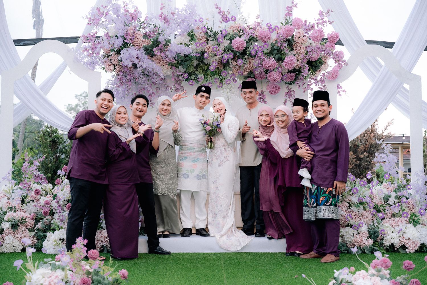 Wedding group photo in purple baju kurung and baju melayu with floral arch backdrop at Malay wedding.