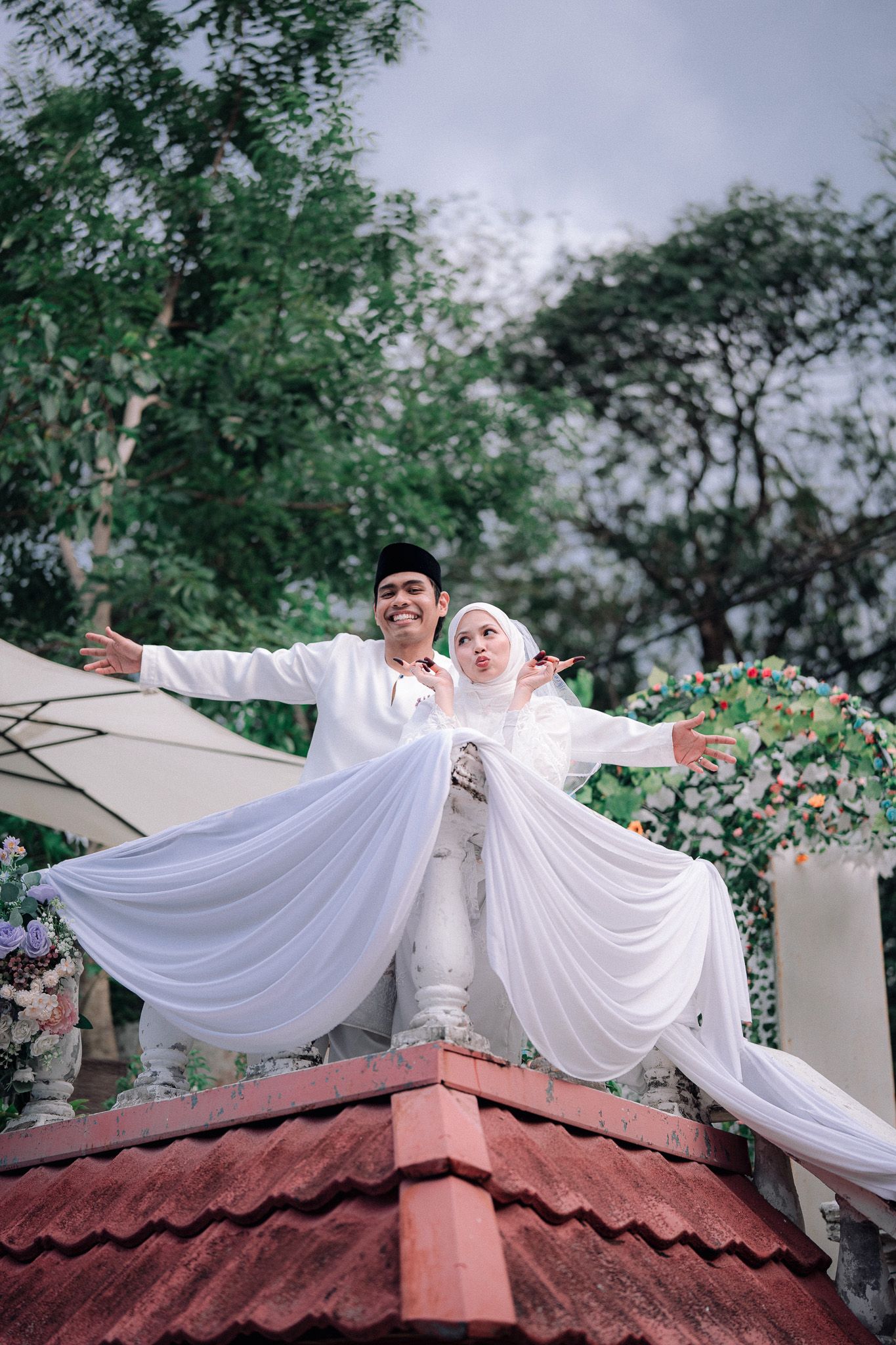 Muslim couple in white wedding attire posing joyfully on rooftop with arms outstretched, surrounded by lush green trees.
