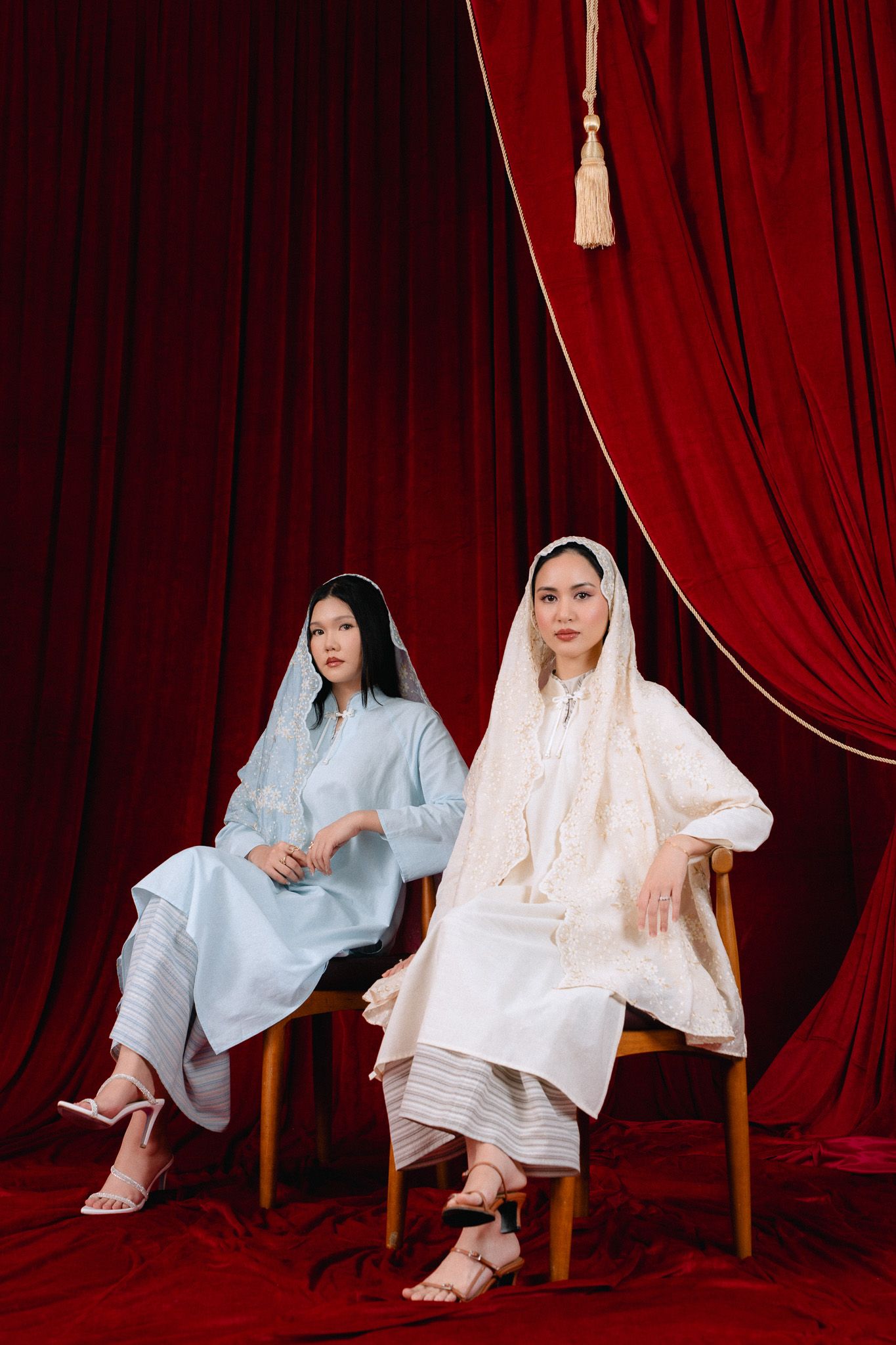 Two women in elegant white and blue traditional outfits with headscarves seated before red velvet curtains.