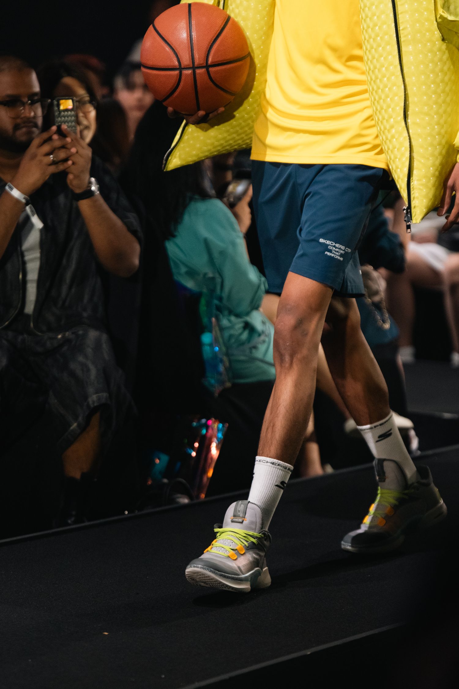 Close-up of basketball and sneakers on a dark stage with people in the background.
