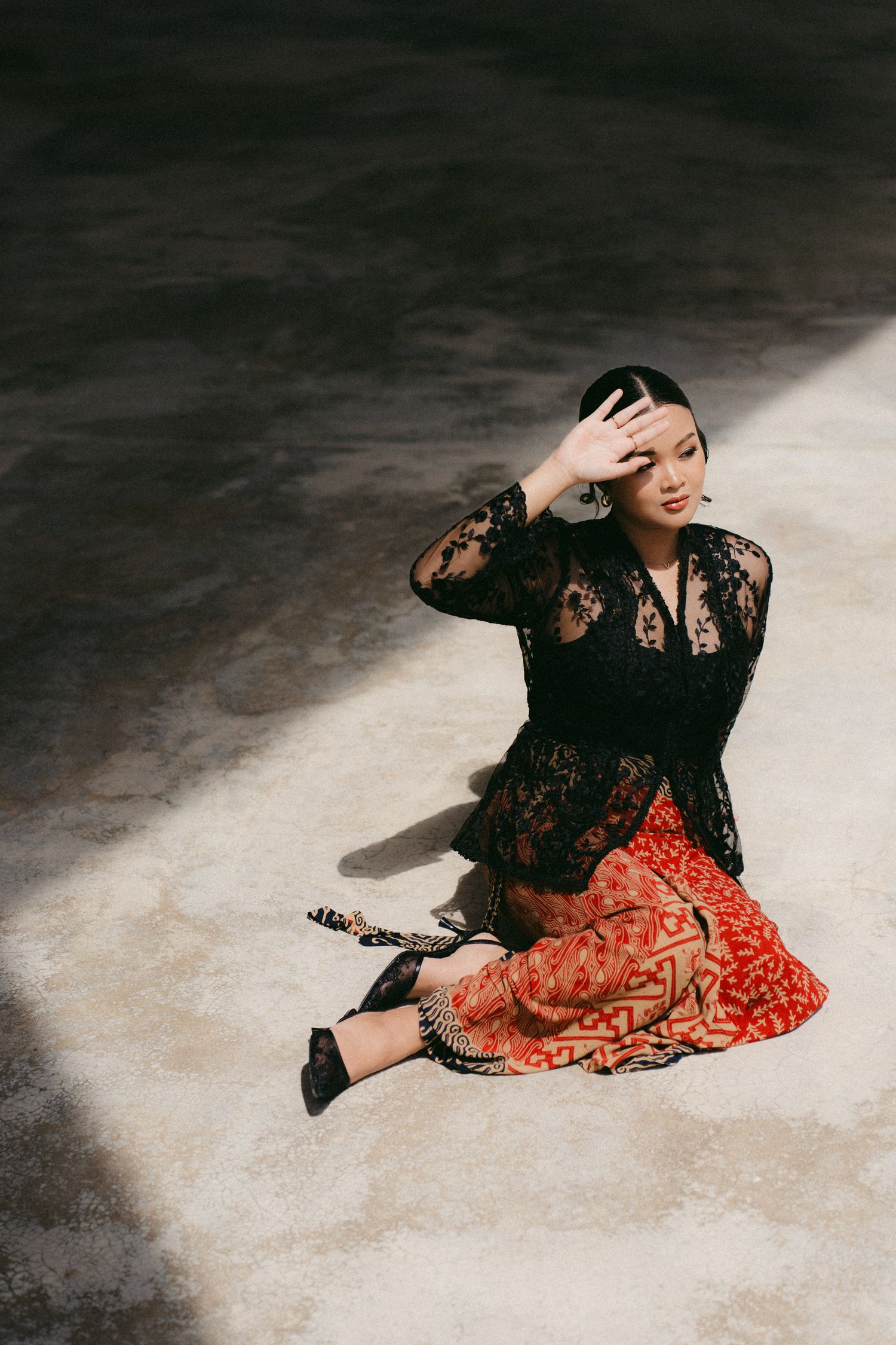 A person in a black lace top and red patterned skirt sits on a concrete floor in dramatic lighting.