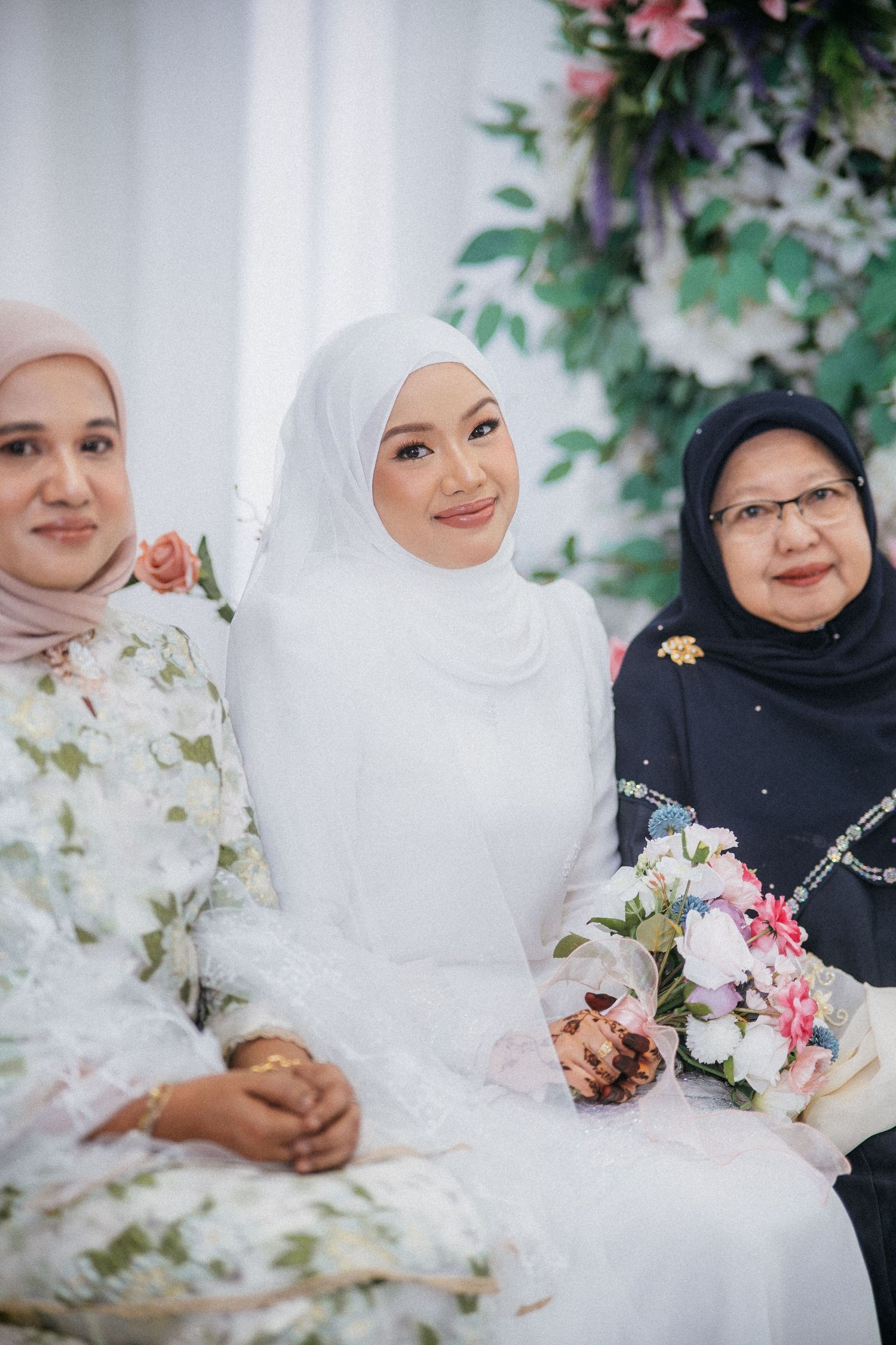 Three women wearing hijabs sit together at a formal event with floral decorations and a white bridal bouquet.