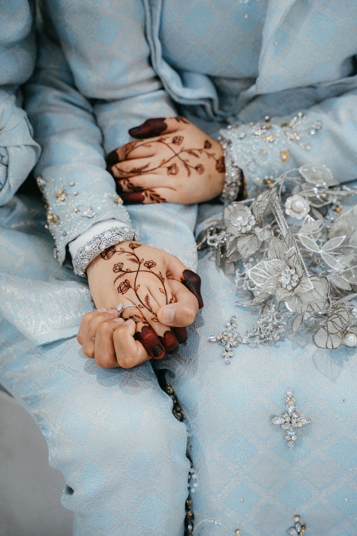 Hands with henna designs rest on light blue fabric with delicate silver beaded floral decorations.