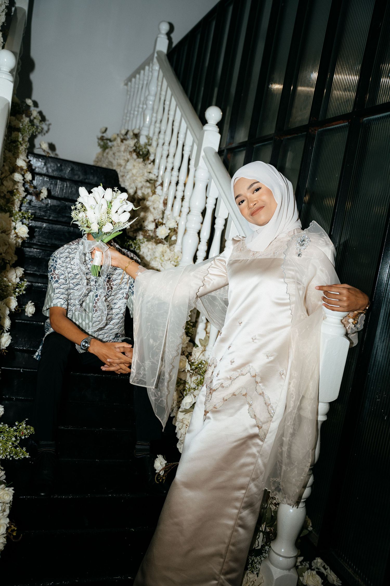 White wedding dress with lace details and flowing sleeves adorns a figure standing by an elegant staircase with floral decor.