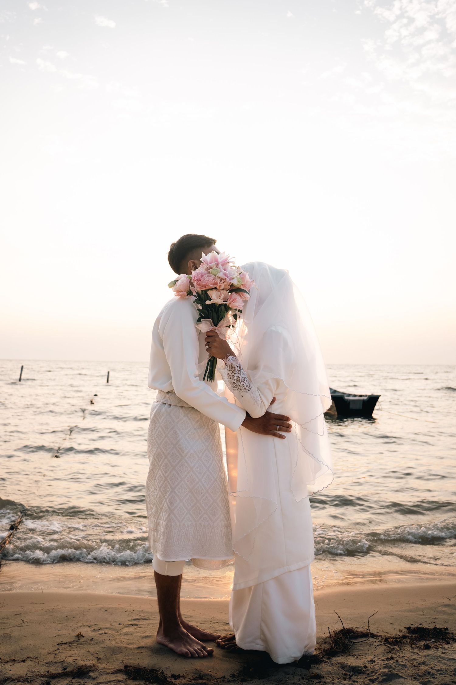 A couple in white attire sharing intimate moments during their seaside wedding photoshoot at sunset.