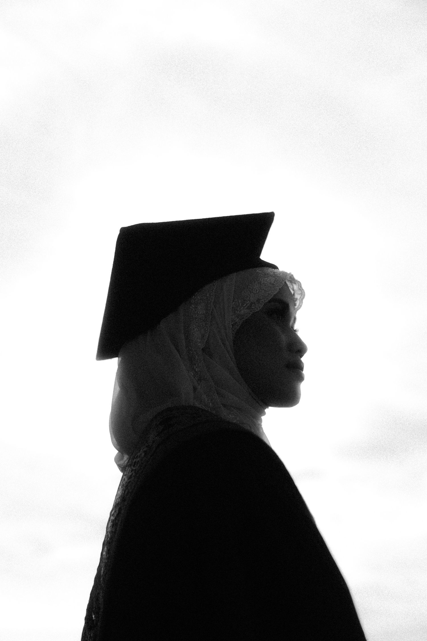 Silhouetted figures in graduation caps and gowns stand against a bright white background in black and white.