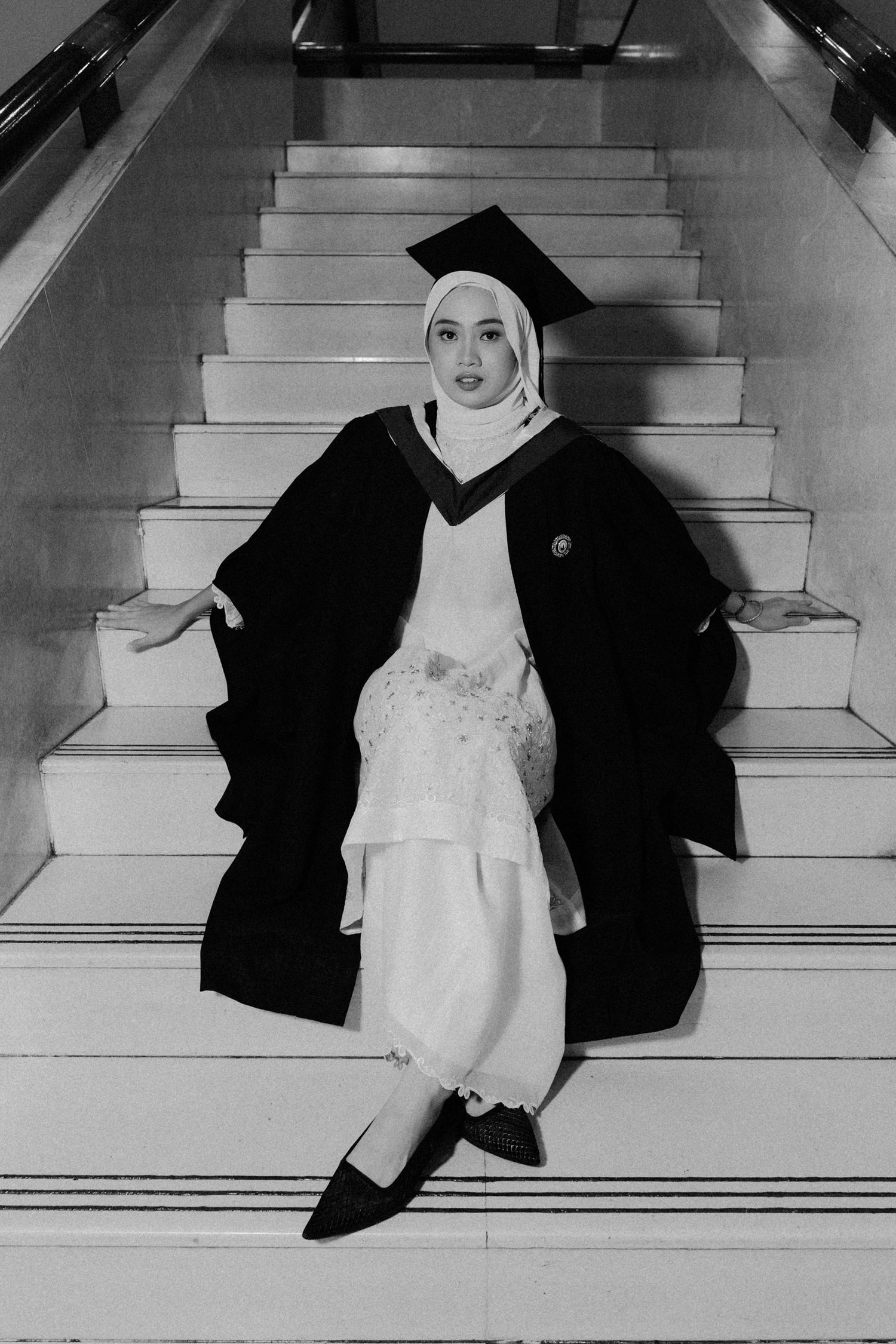 A series of black and white photos showing a person in a hijab and graduation gown posing on stairs.