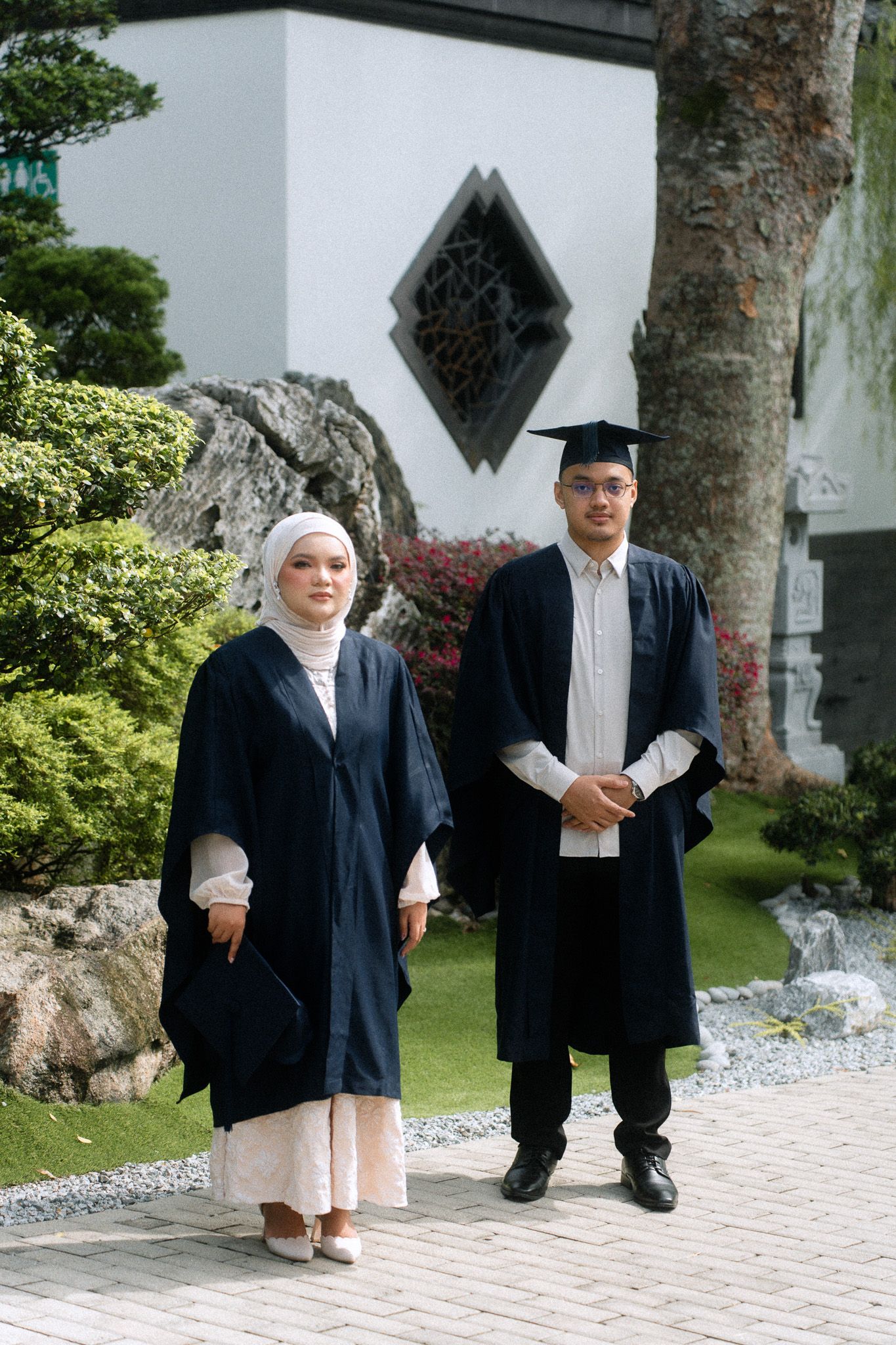 Two graduates in navy gowns pose outdoors, woman in hijab holds cap, man wears mortarboard, garden setting behind them.