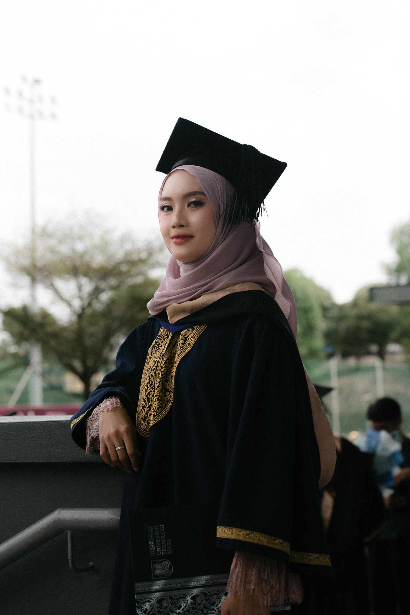 Young Muslim woman in black graduation gown and mortarboard cap with hijab smiling outdoors at commencement ceremony.