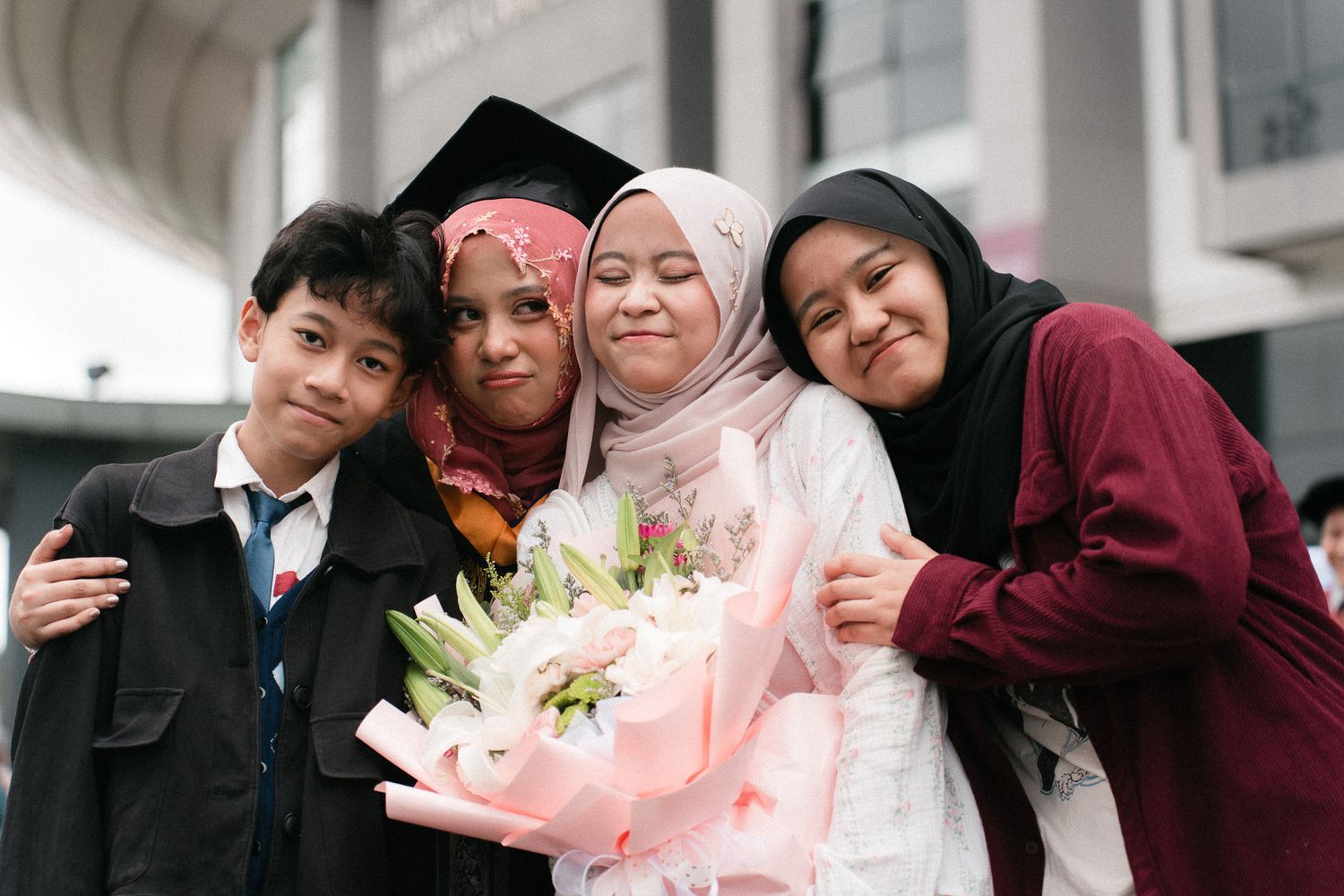 Graduate in cap and hijab celebrates with family holding bouquet of white lilies at graduation ceremony.