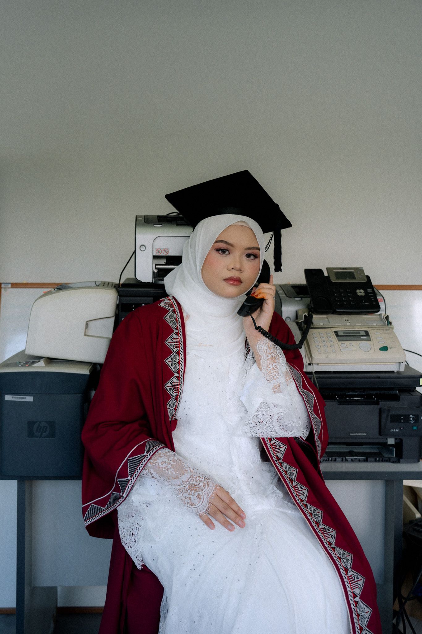Muslim woman in white hijab and red graduation gown sitting by office printers holding phone.