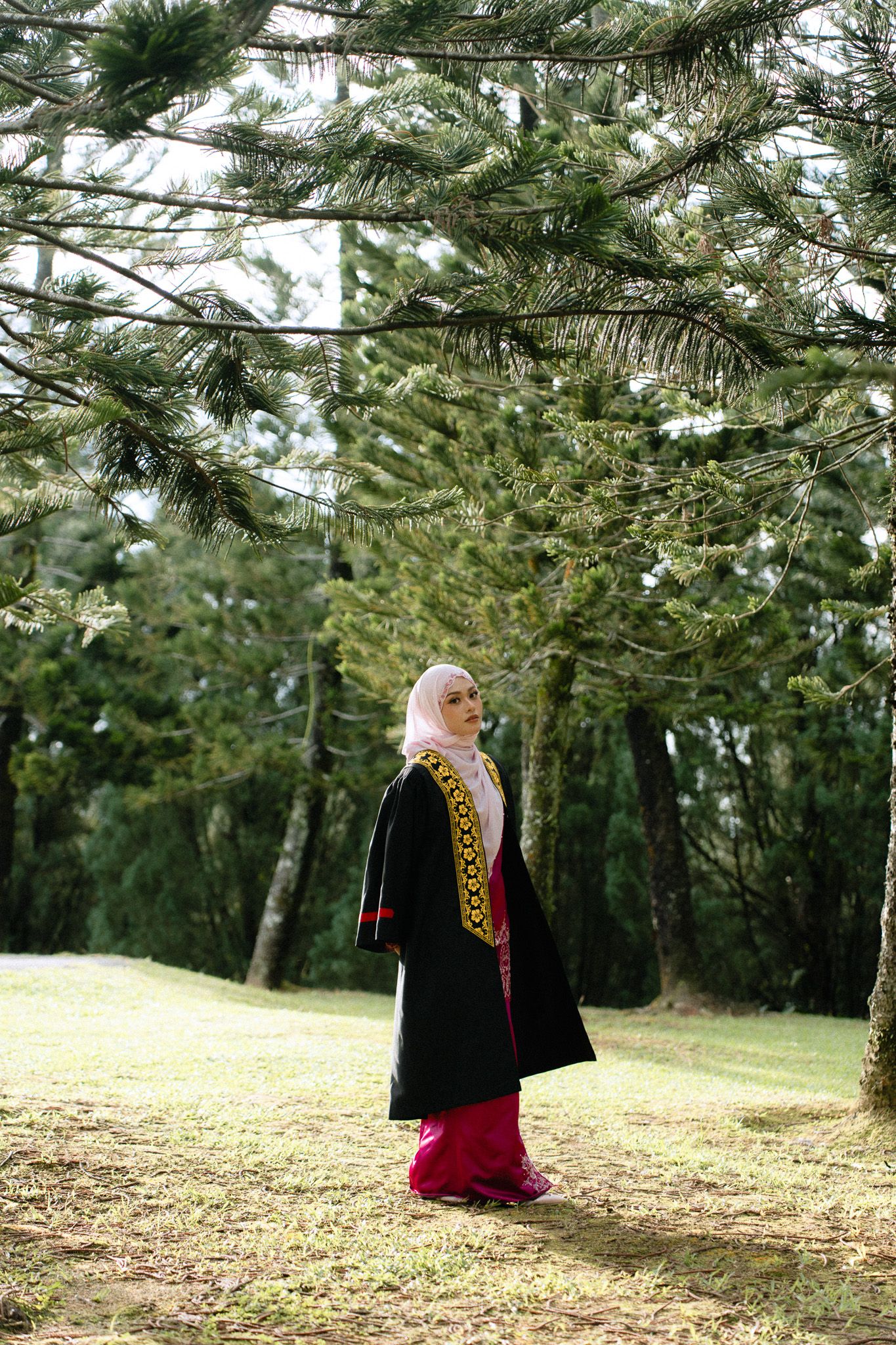 Muslim woman in black abaya and pink hijab standing gracefully among green pine trees in a serene outdoor park.