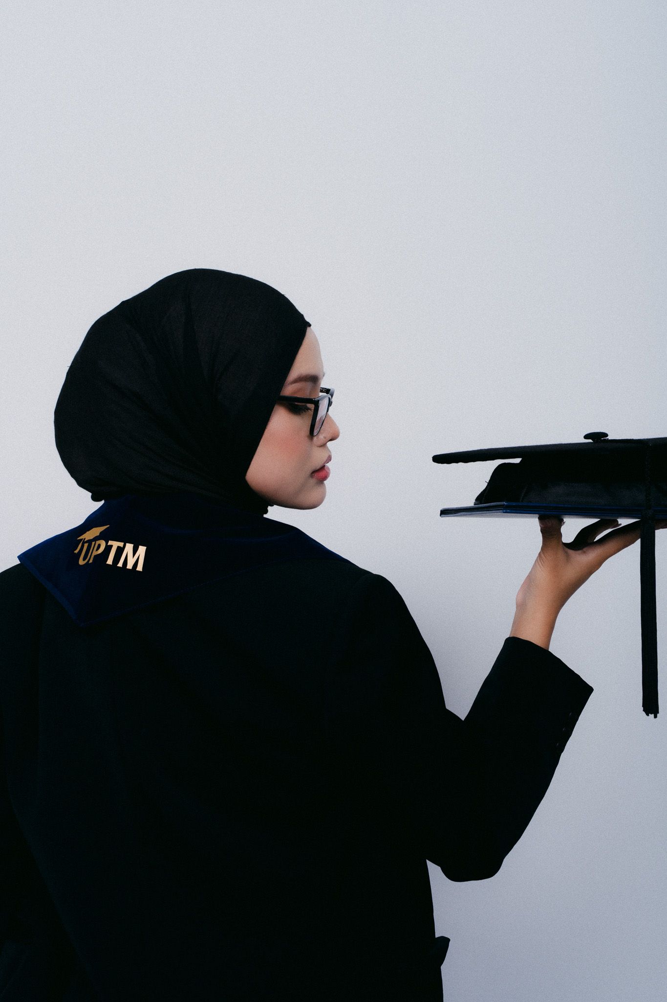 Muslim woman in black hijab and graduation gown holding mortarboard cap, side profile view against white background.