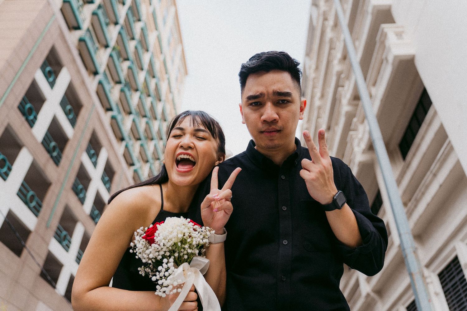 A couple in formal attire making peace signs and smiling in front of an urban building.
