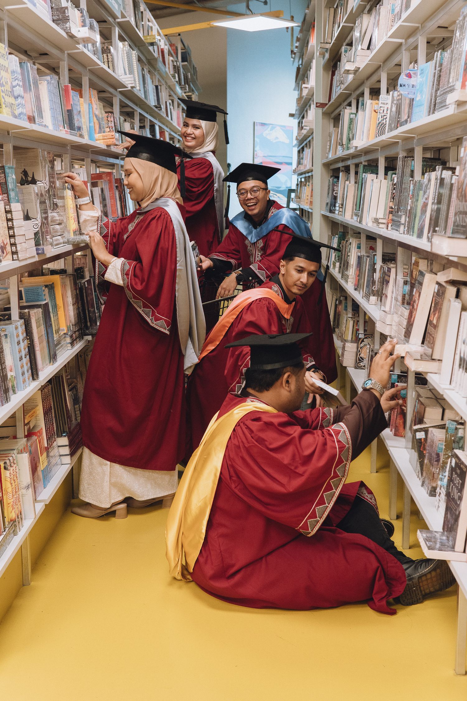 Graduates in red and gold regalia pose creatively between library bookshelves.