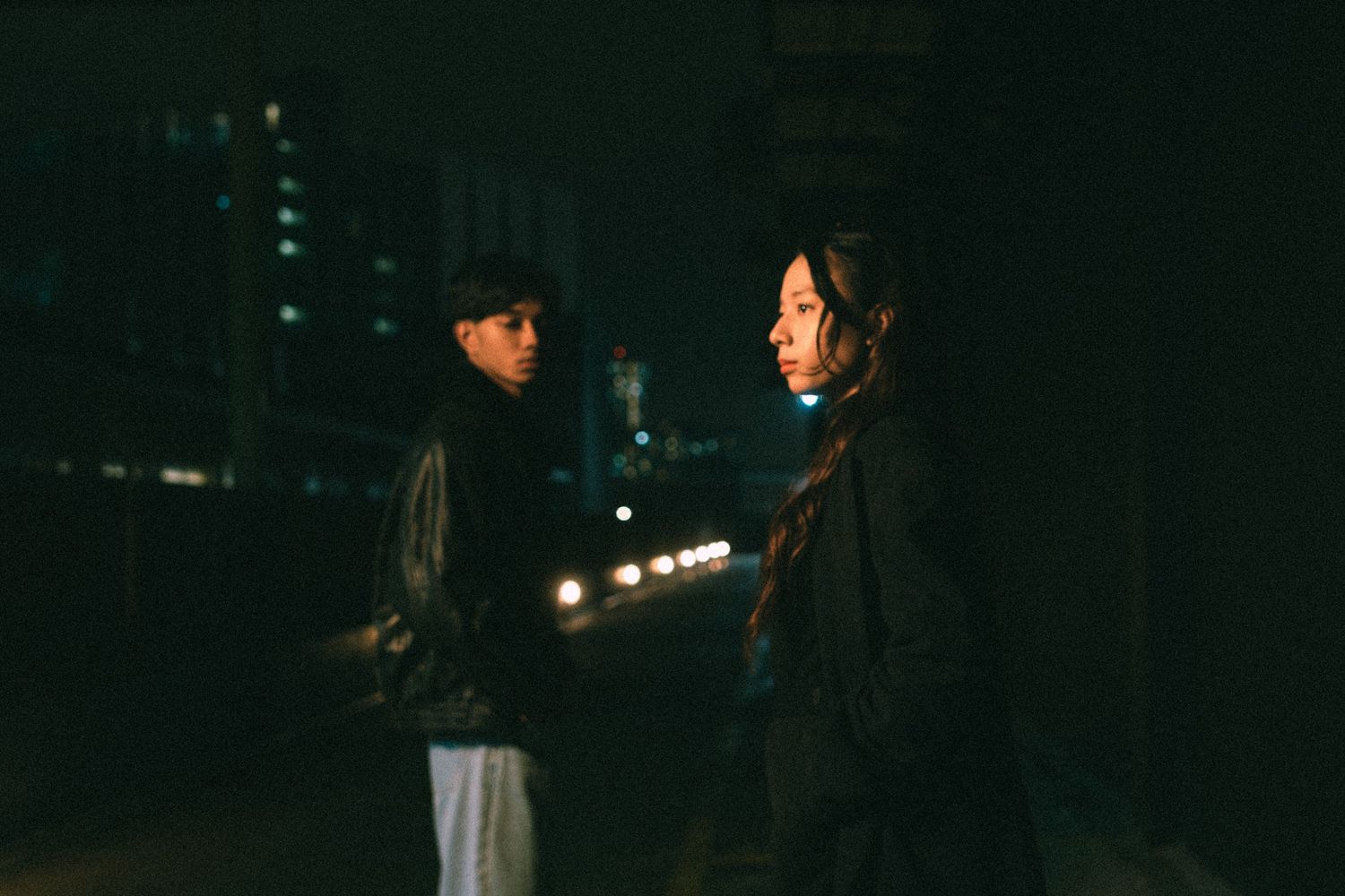 Two people having a conversation on a dark city street illuminated by ambient lights.