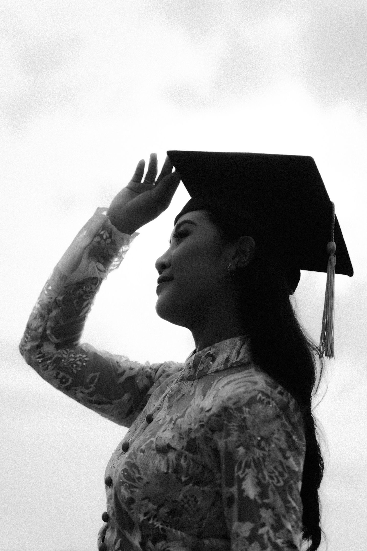 A person in graduation attire looks up at the sky in a black and white silhouette photo series.