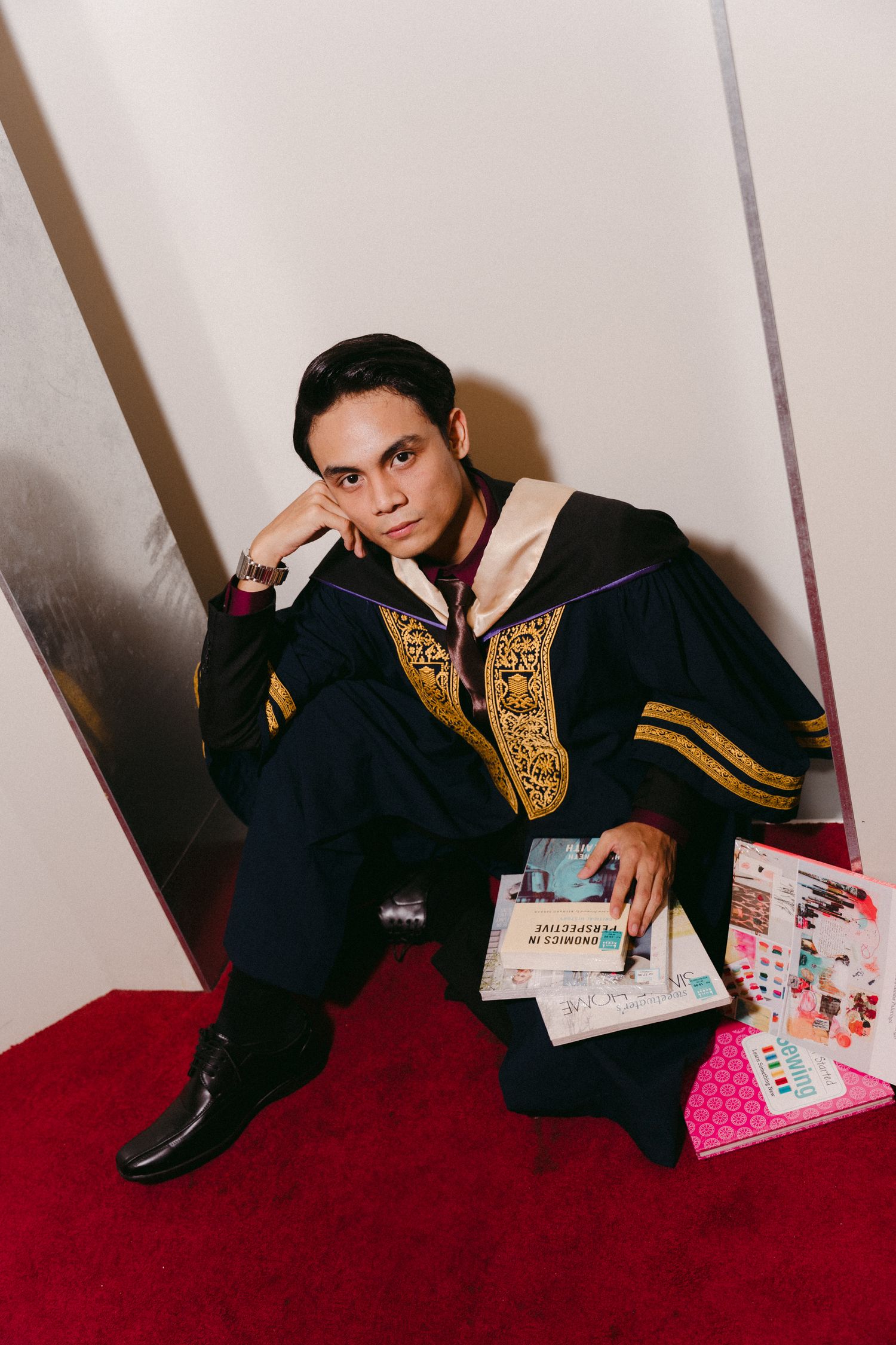 Graduate in black and gold embroidered robe posing with books on red carpet.