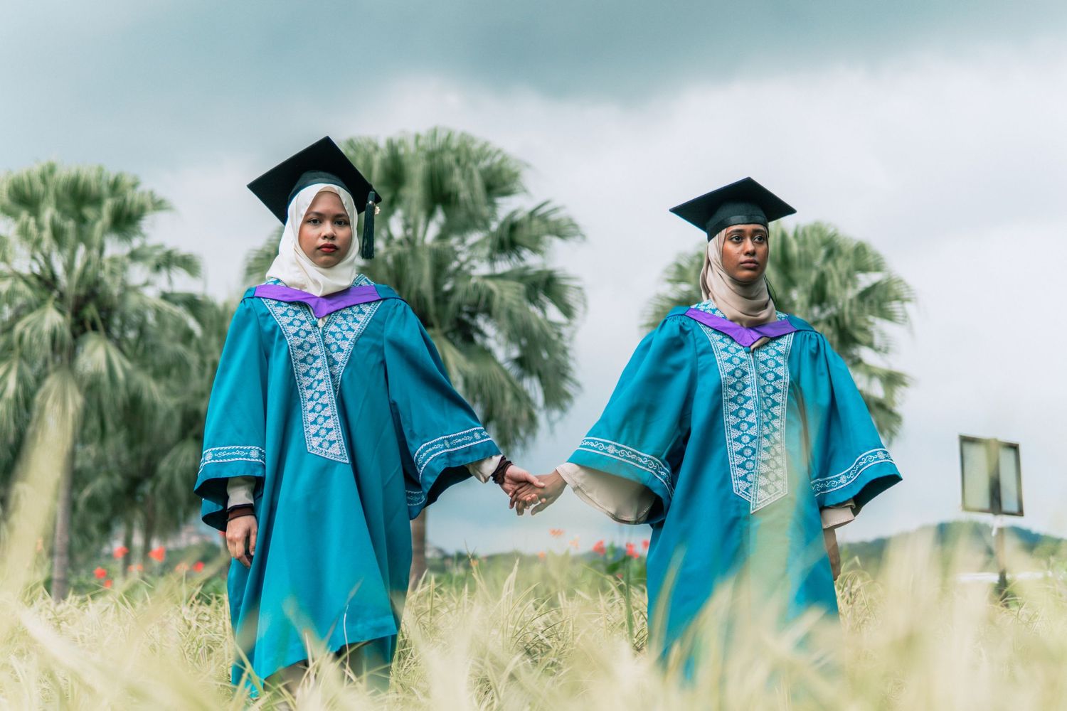 Two graduates in turquoise graduation gowns walking through grass field.