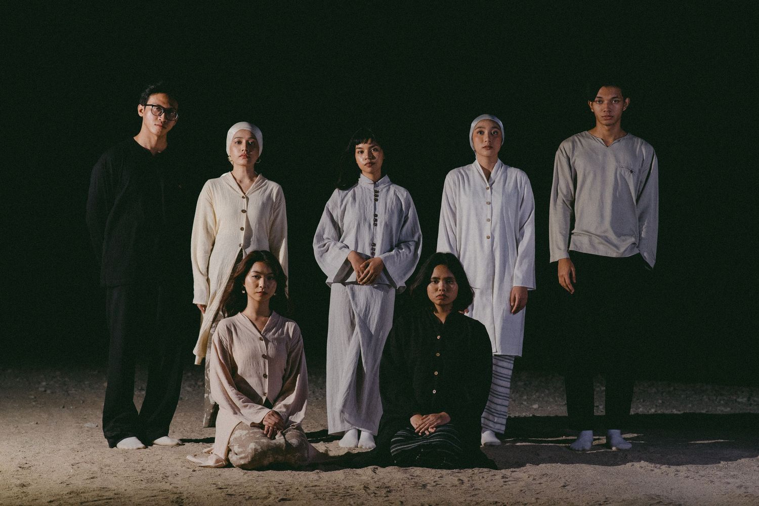 Group portrait of seven people wearing black and white clothing in dramatic lighting.