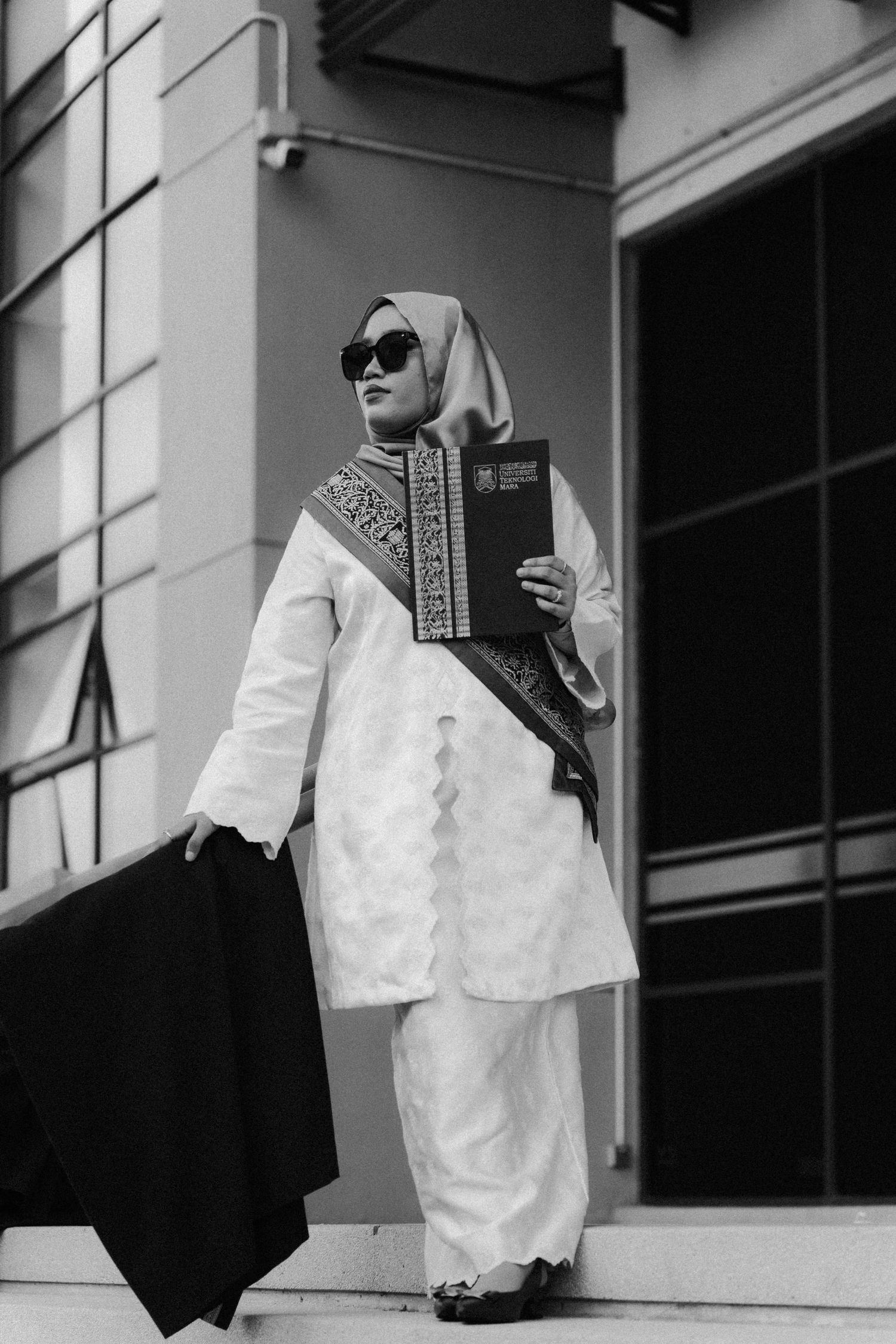 Person in white modest fashion attire with hijab and sunglasses poses on steps in black and white photography.