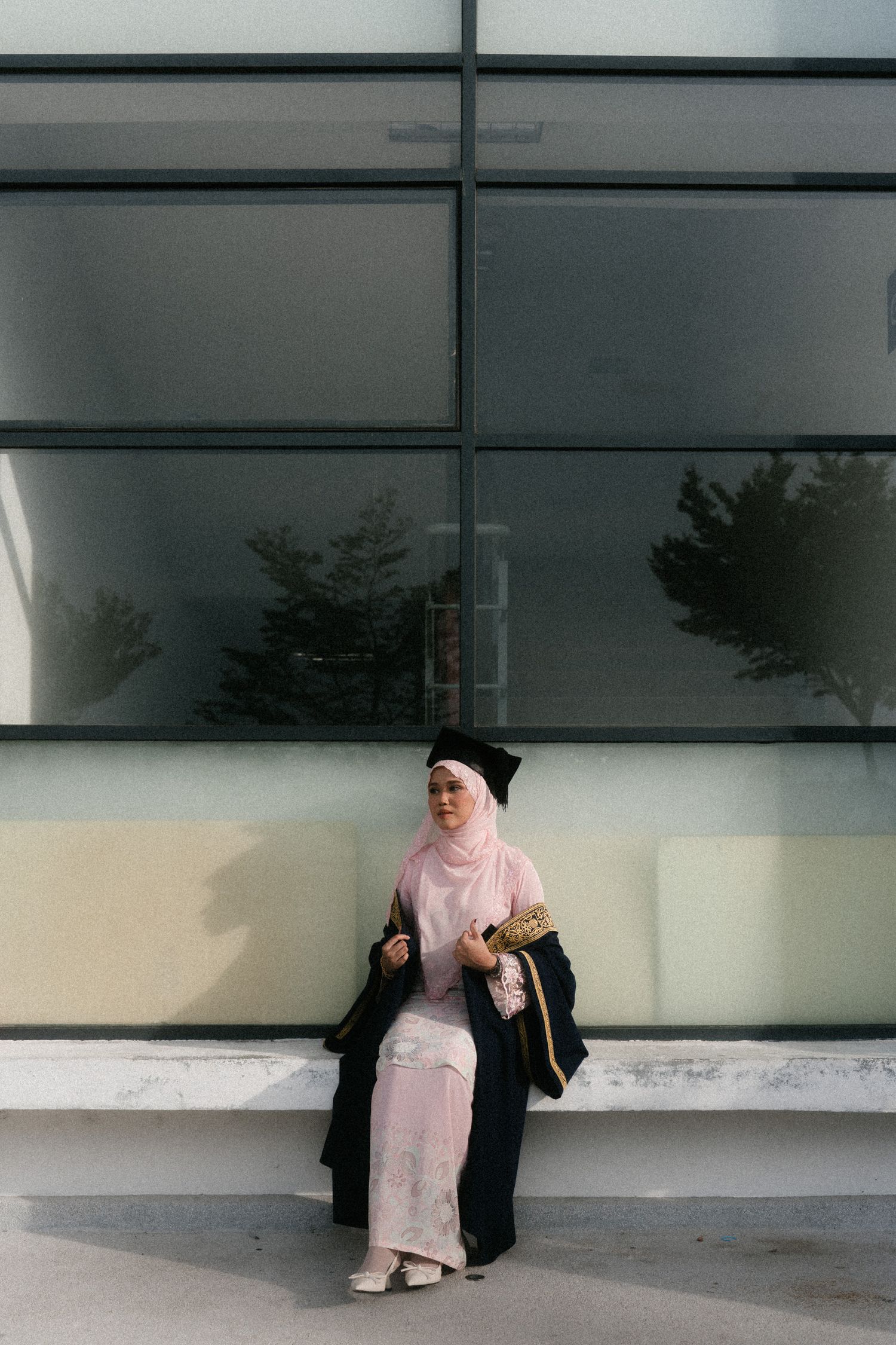 Graduate in black academic regalia and pink hijab sits against modern glass building exterior.