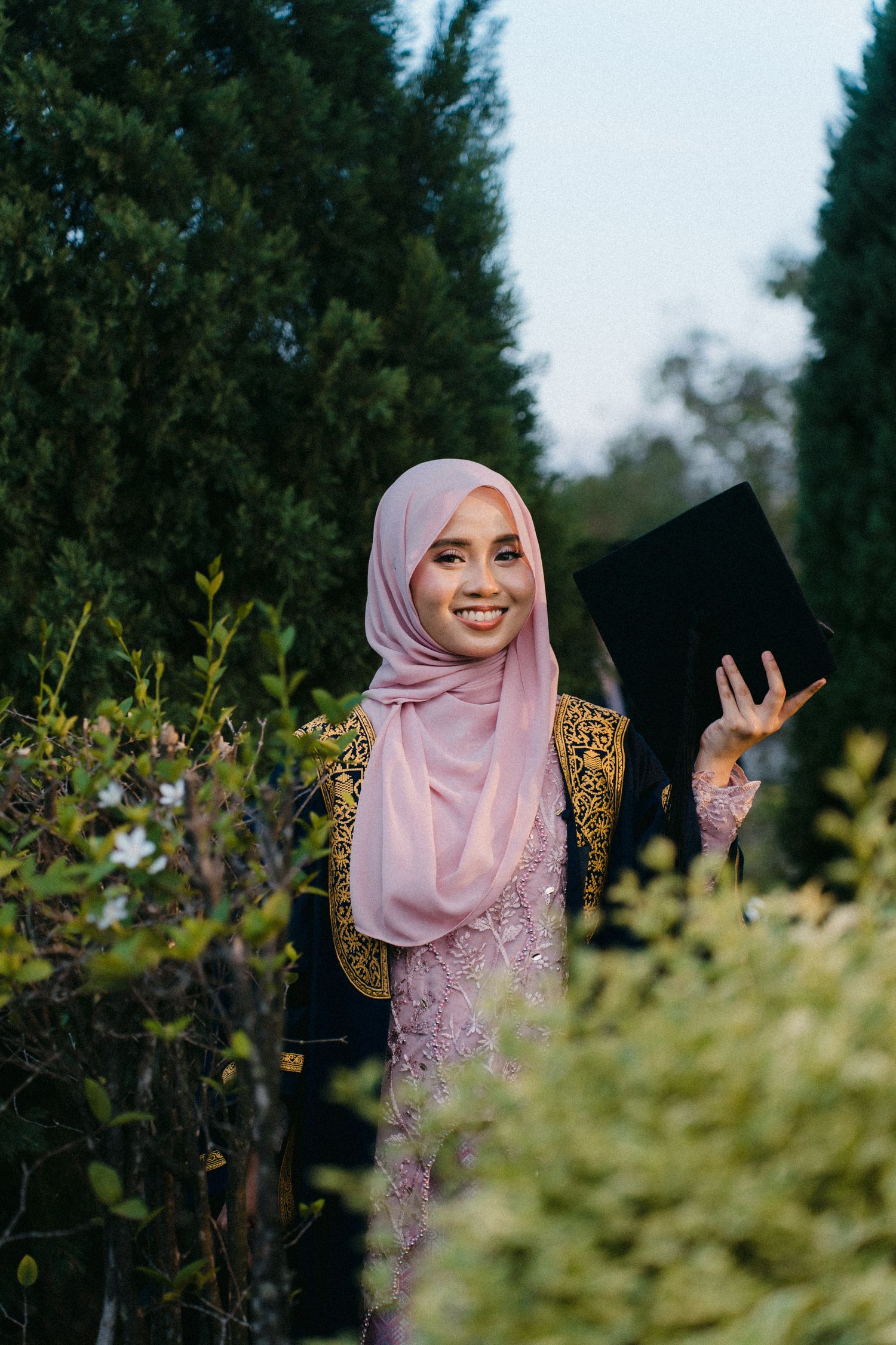 A person in a pink hijab and floral dress poses joyfully outdoors among green foliage and trees.