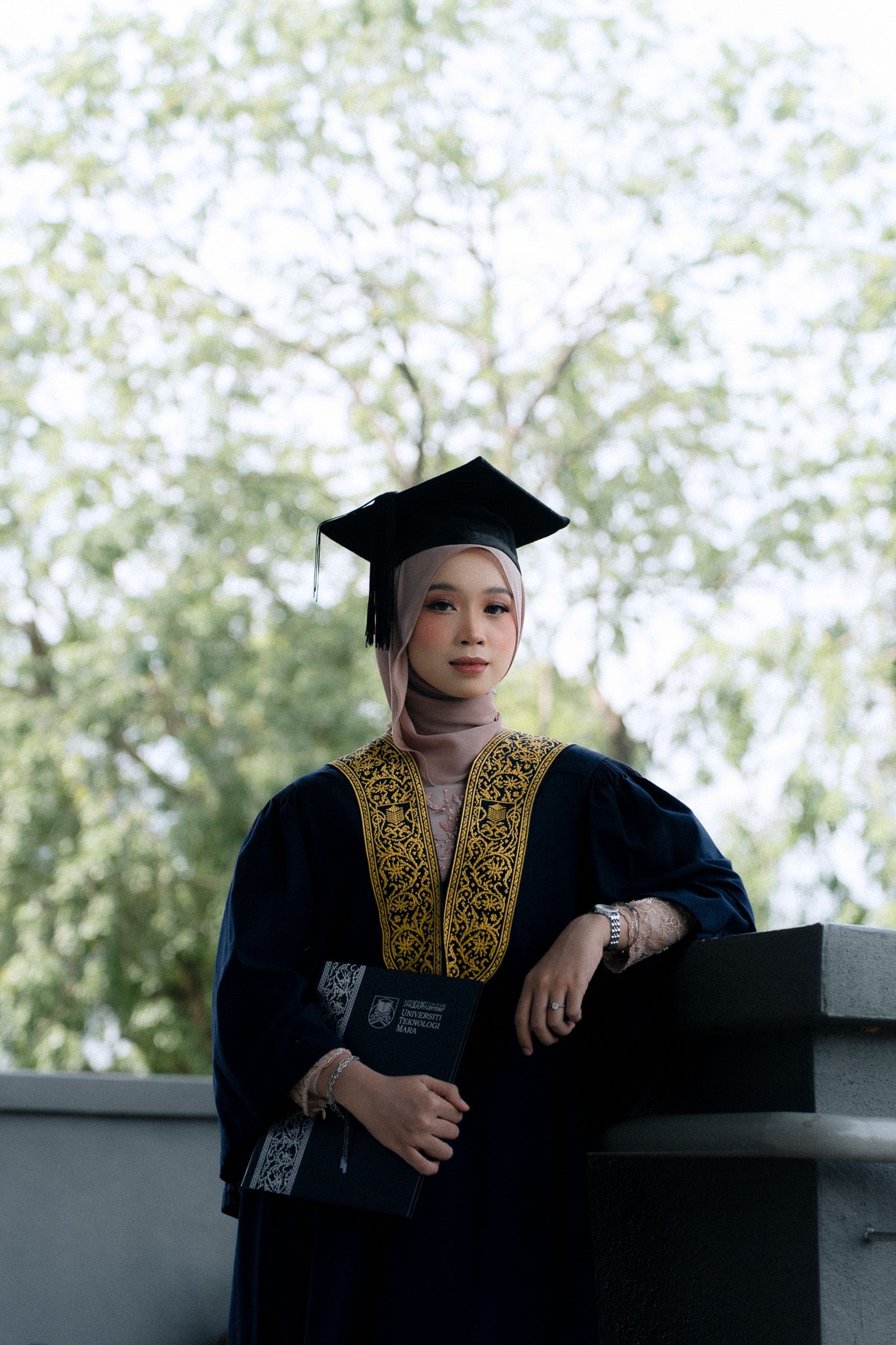 A young woman in graduation gown, mortarboard cap, and hijab holds a diploma while posing outdoors.