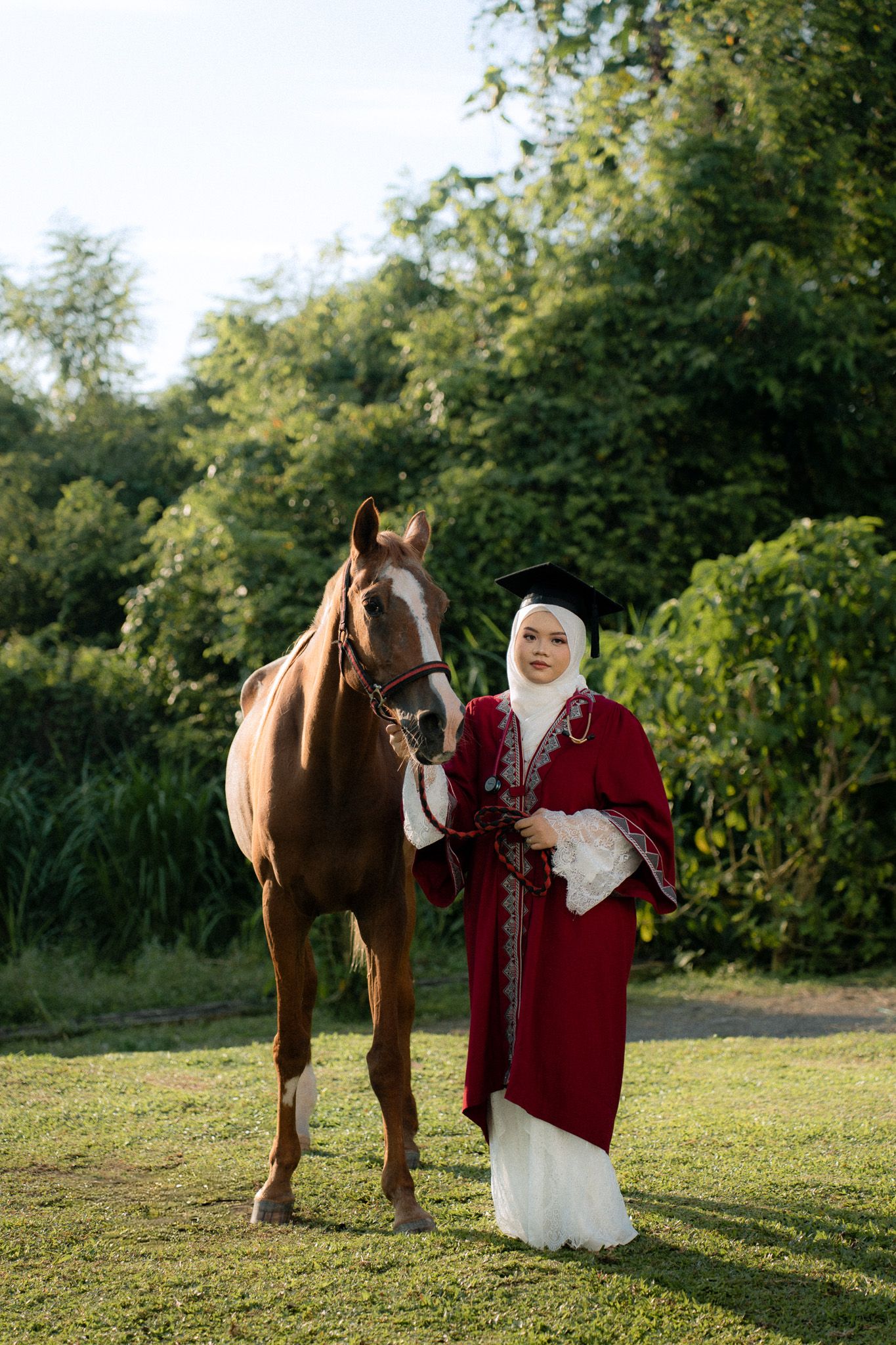 Muslim woman in red graduation gown and hijab stands proudly beside a brown horse on green grass outdoors.