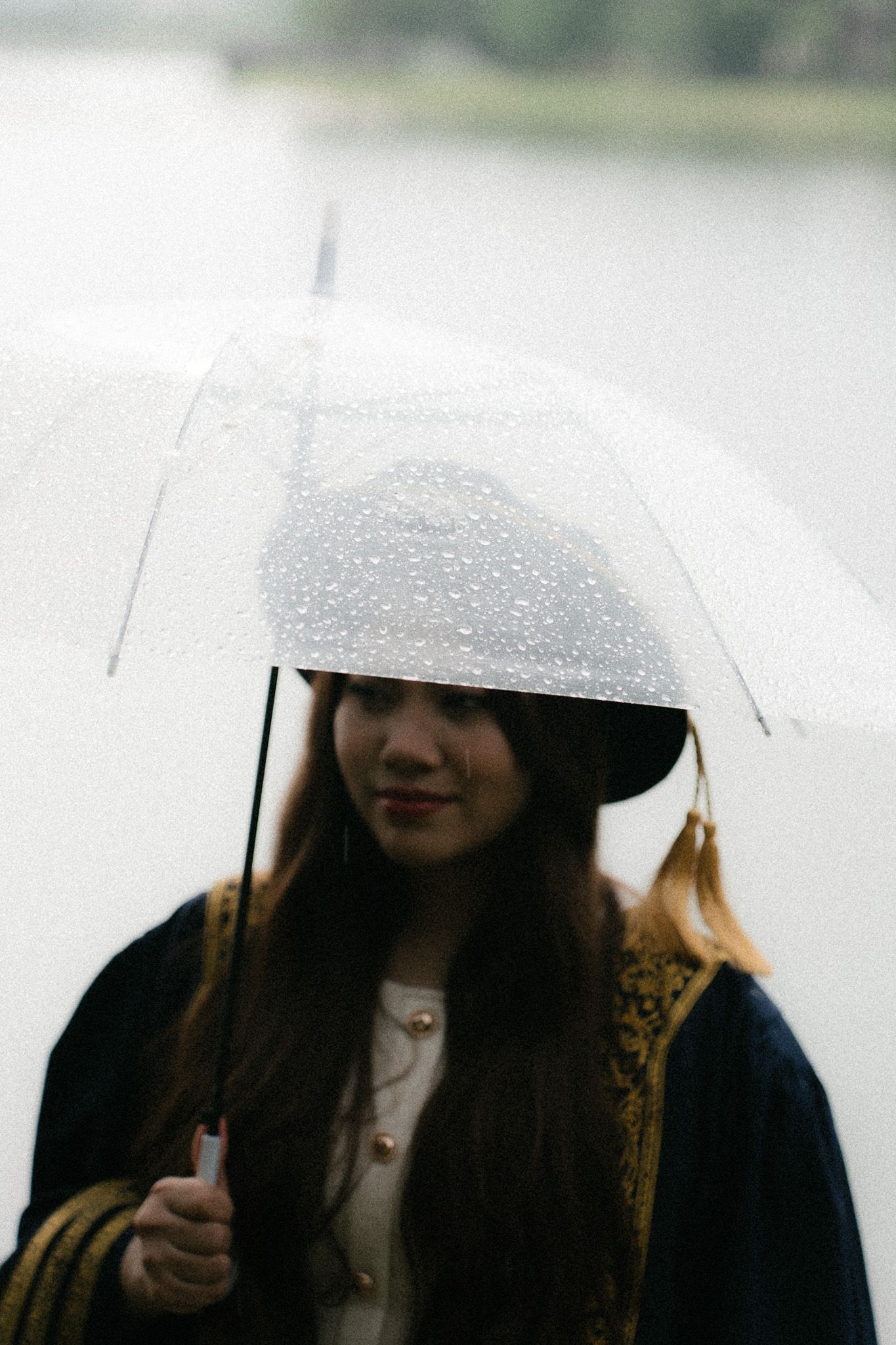 Young woman holding a clear umbrella with raindrops in the rain, wearing a dark coat and hat outdoors.