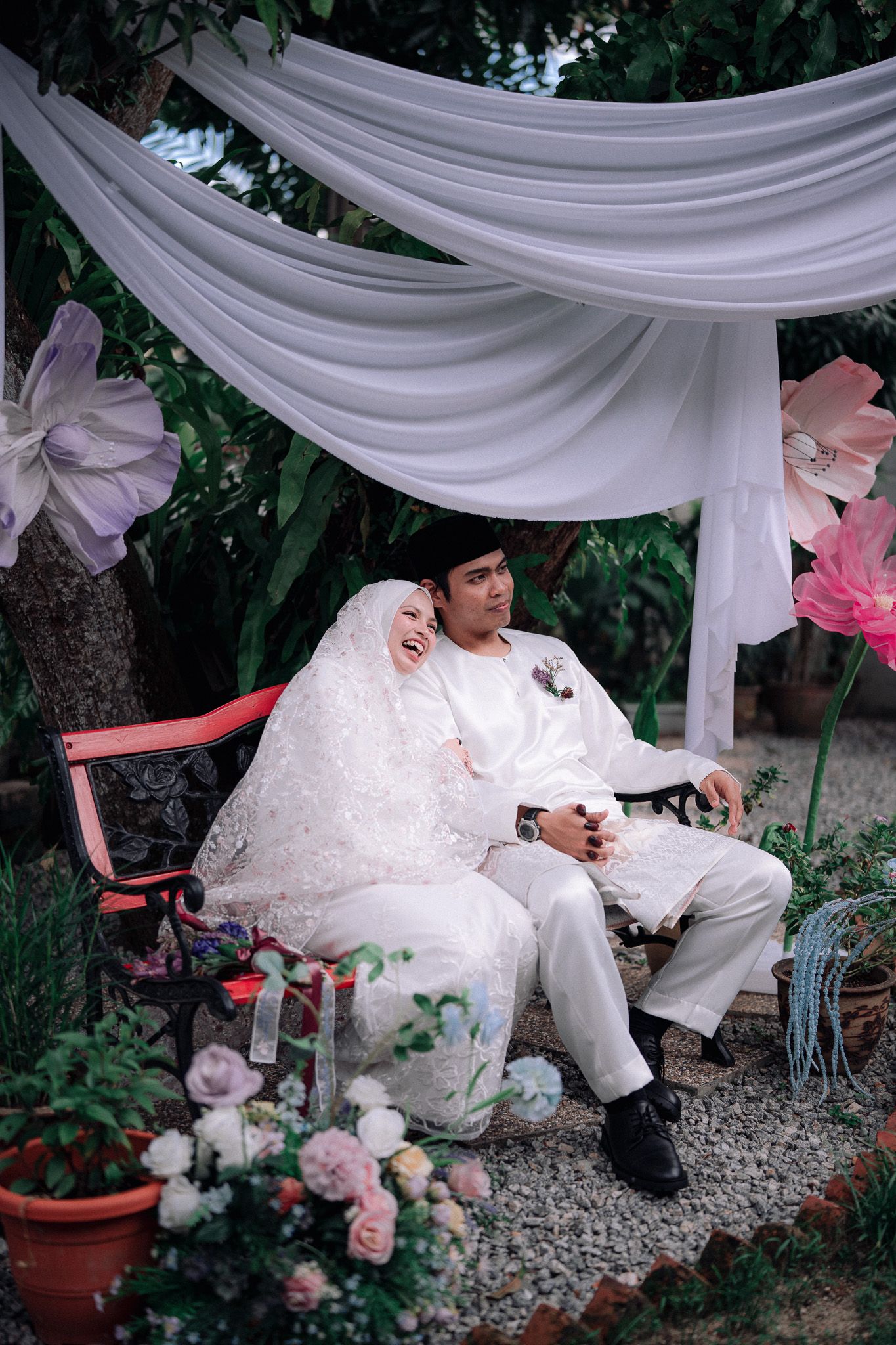 Muslim couple in white wedding attire sitting on a bench under draped fabric canopy surrounded by flowers in a garden.